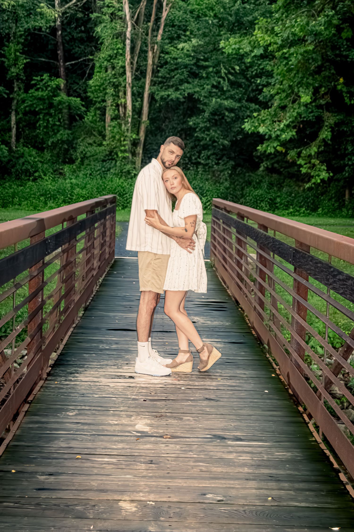 A couple in matching white outfits embraces on a wooden bridge surrounded by lush green forest.