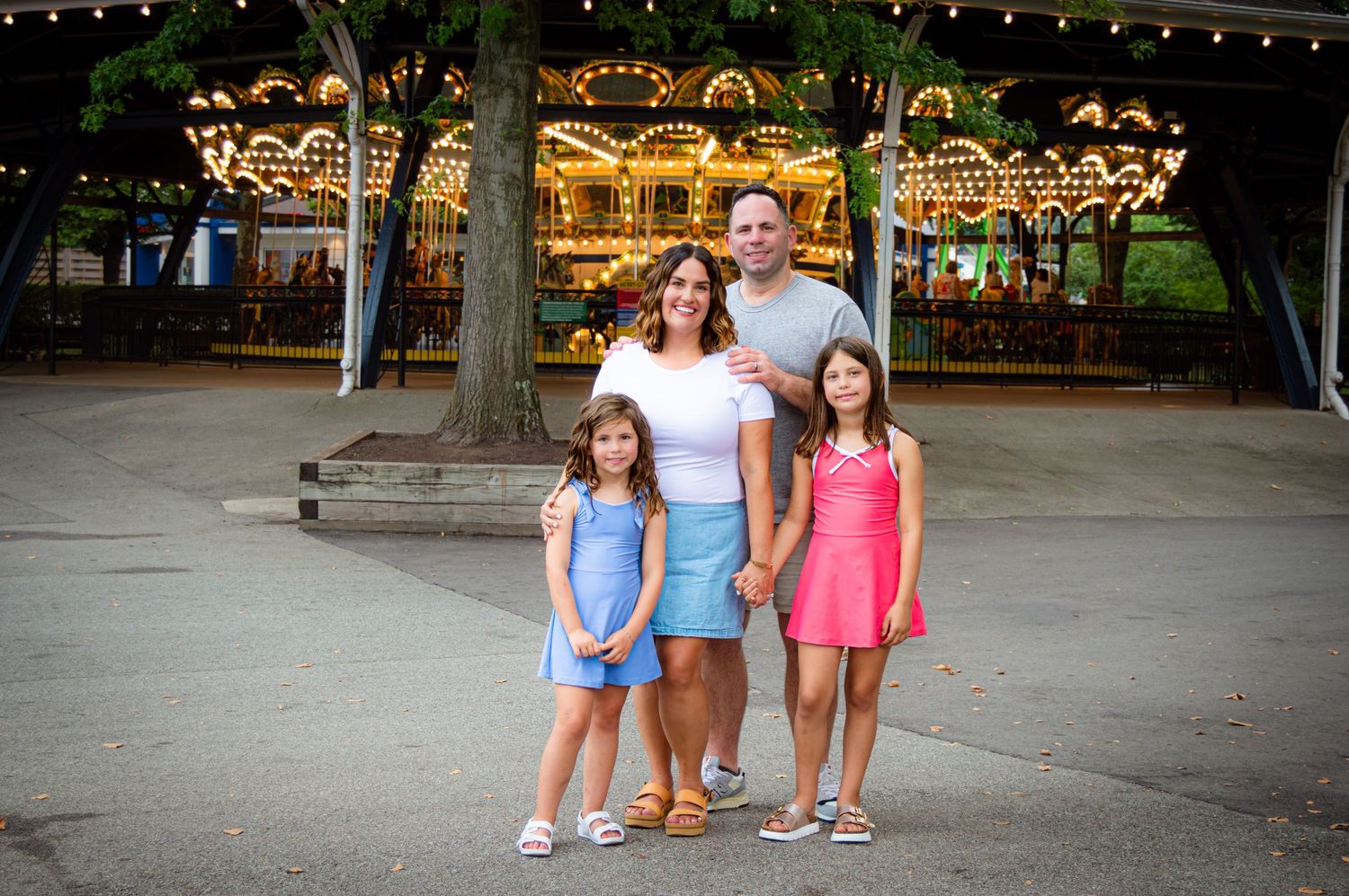 Family of four smiling together near a lit carousel at dusk in amusement park.