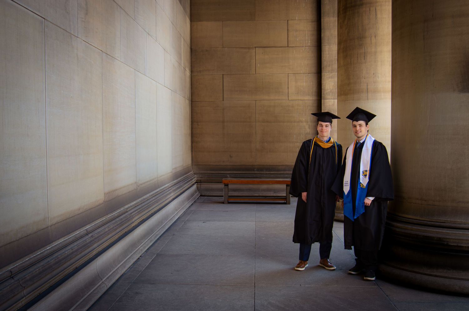 Two graduates in black caps and gowns pose between stone columns for a graduation portrait.