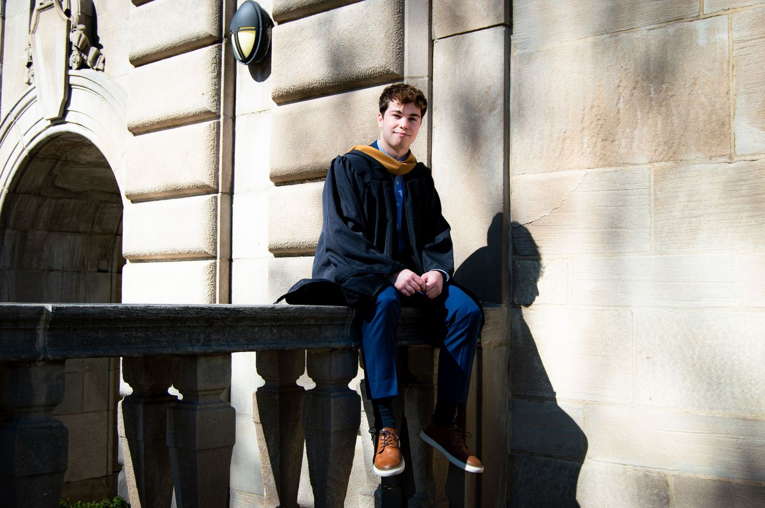 Graduate in navy cap and gown sitting on stone railing outdoors.