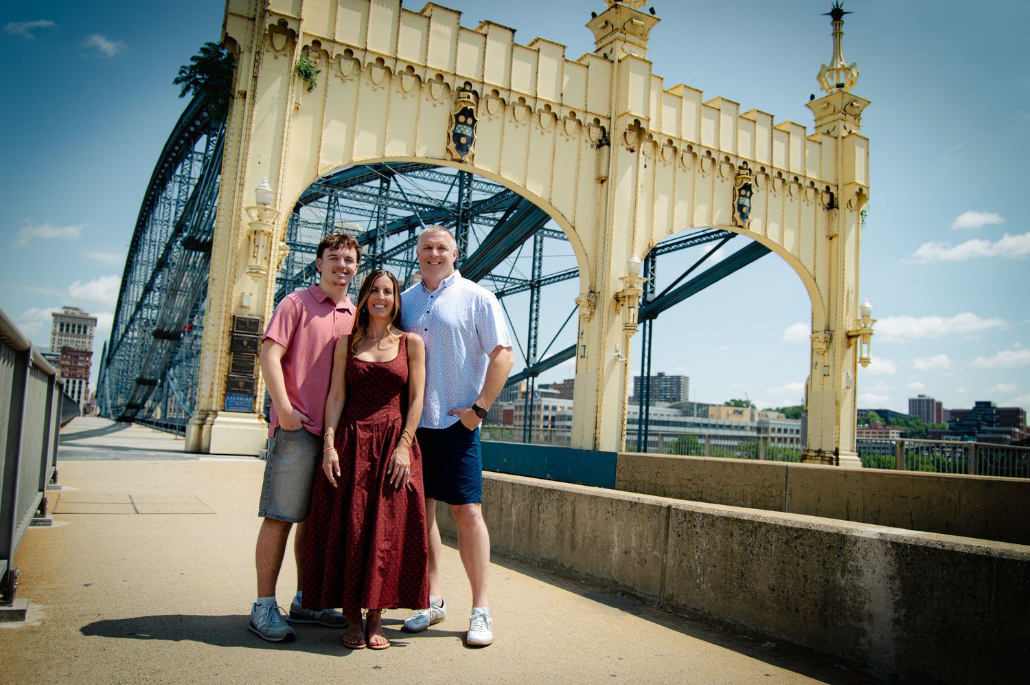 Family of three posing on a historic bridge with steel arch structure on a sunny day.