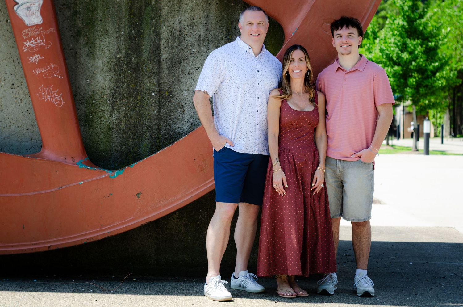 Three people smile together outdoors beside a large red abstract sculpture on a sunny day.