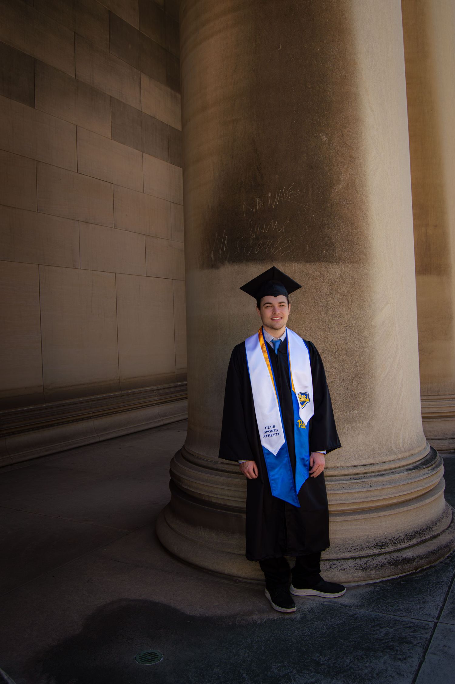 Graduate in cap and gown with blue and white stoles standing beside large stone column.