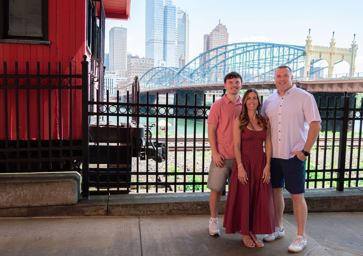 Family of three posing near a red caboose with Pittsburgh skyline in the background.