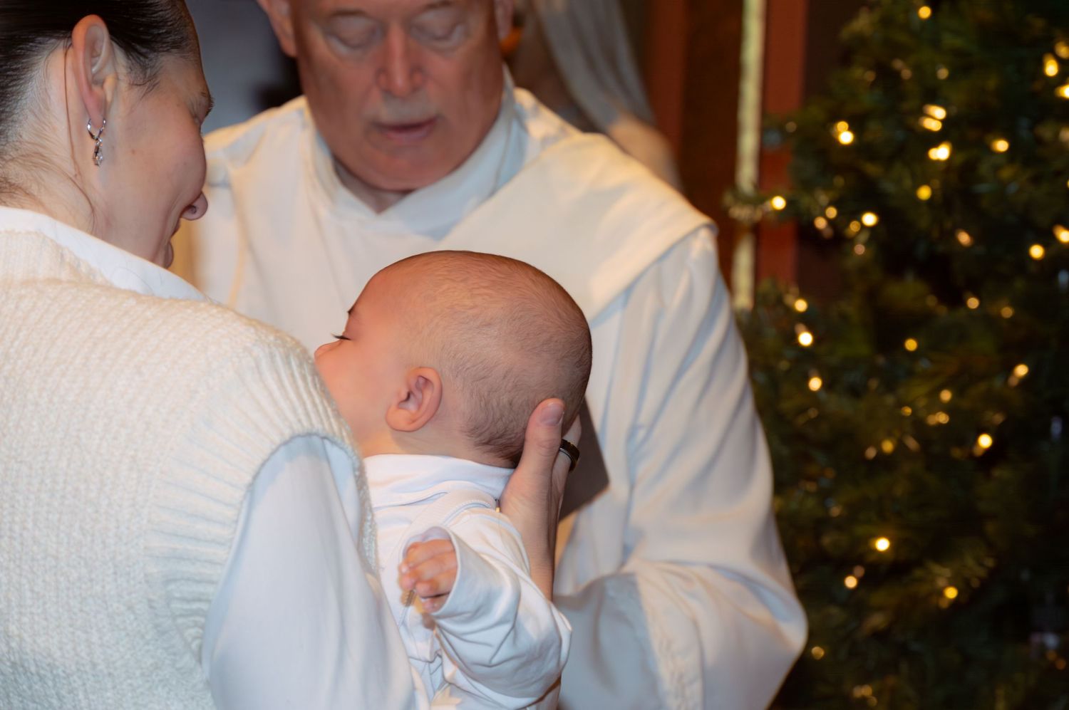 A baby in white clothing is held during what appears to be a Christmas gathering with a decorated tree visible.