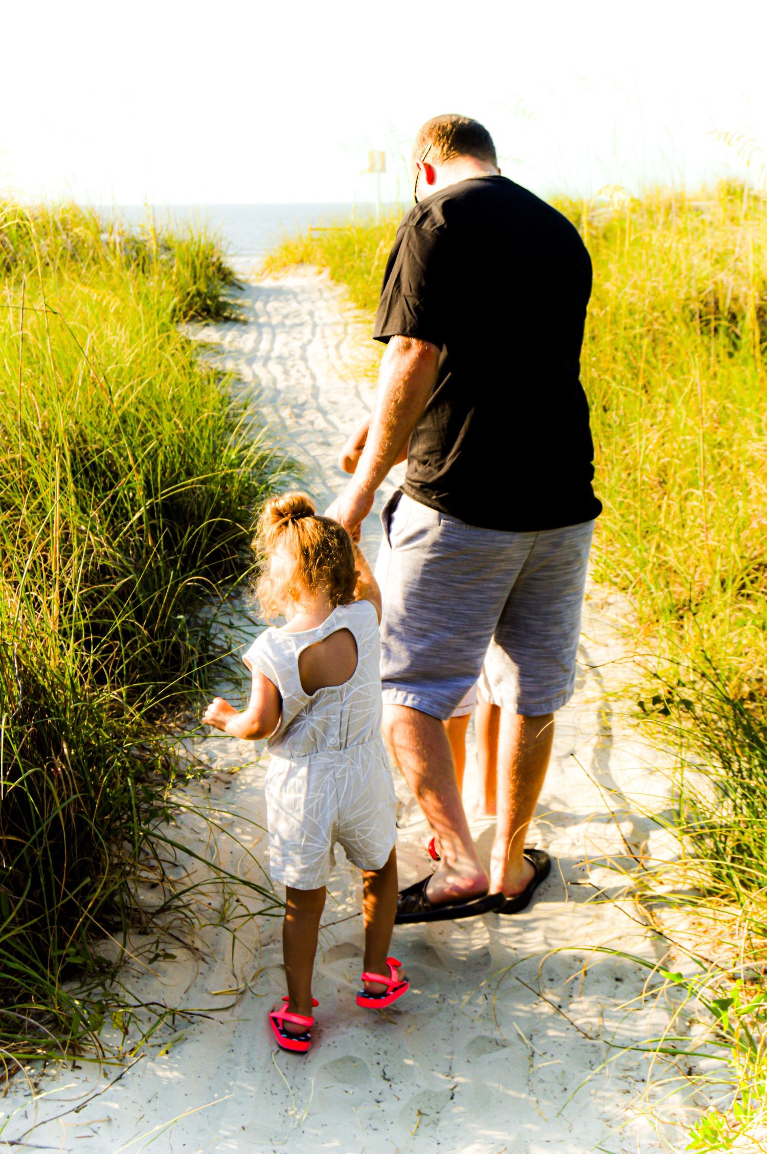 A person and small child walk along a sandy beach path through tall grass on a sunny day.
