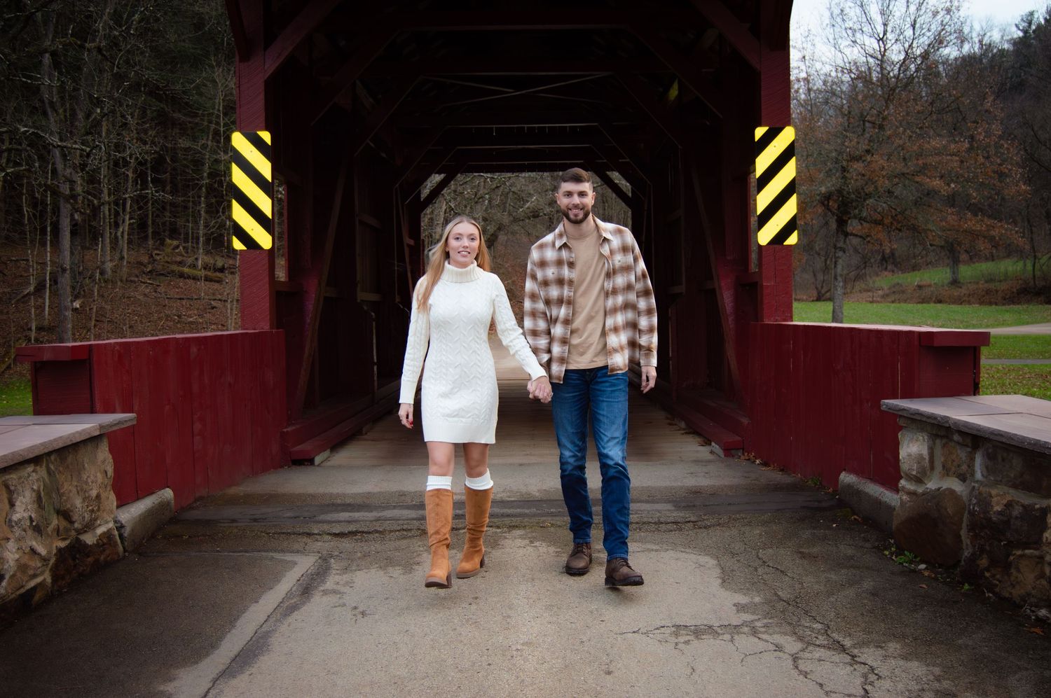 A couple walks hand in hand through a rustic covered bridge with red wooden walls.