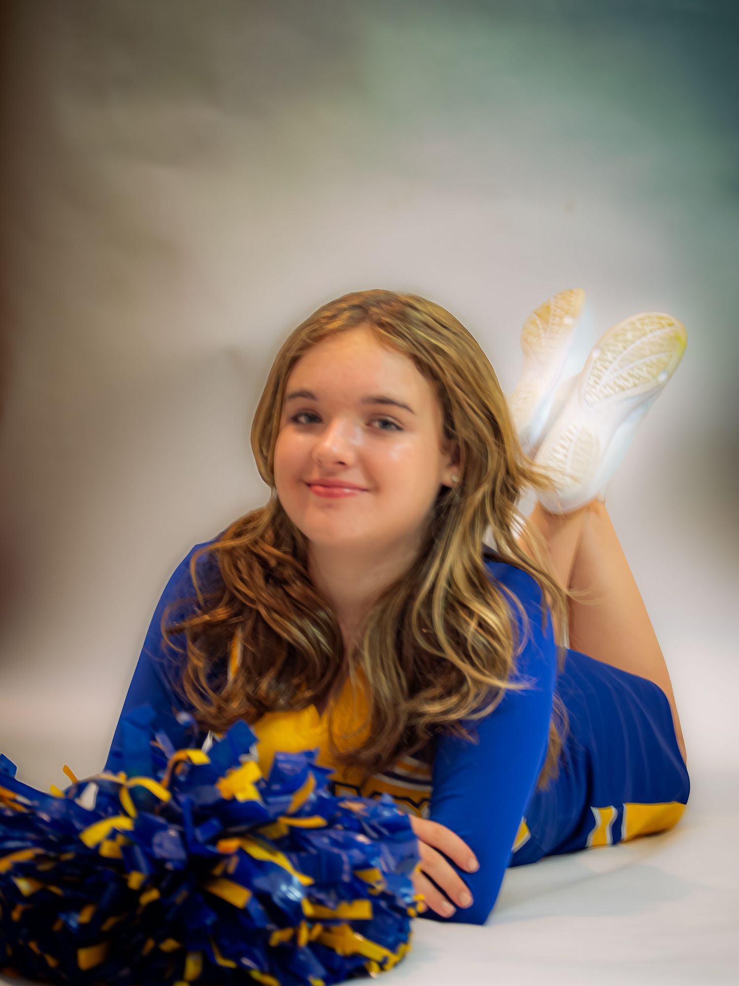 A cheerleader in blue and yellow uniform poses playfully on the floor with pom poms.