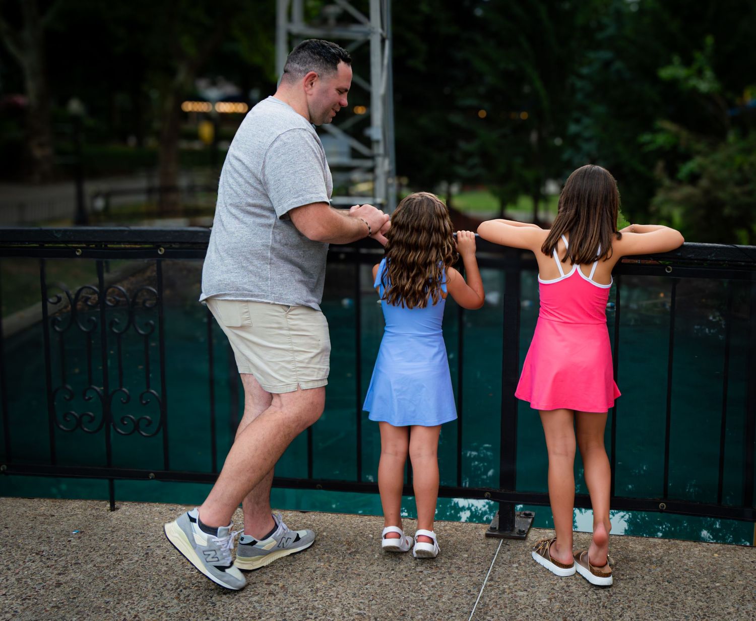A man and two young girls in colorful dresses look over a railing at an outdoor park.