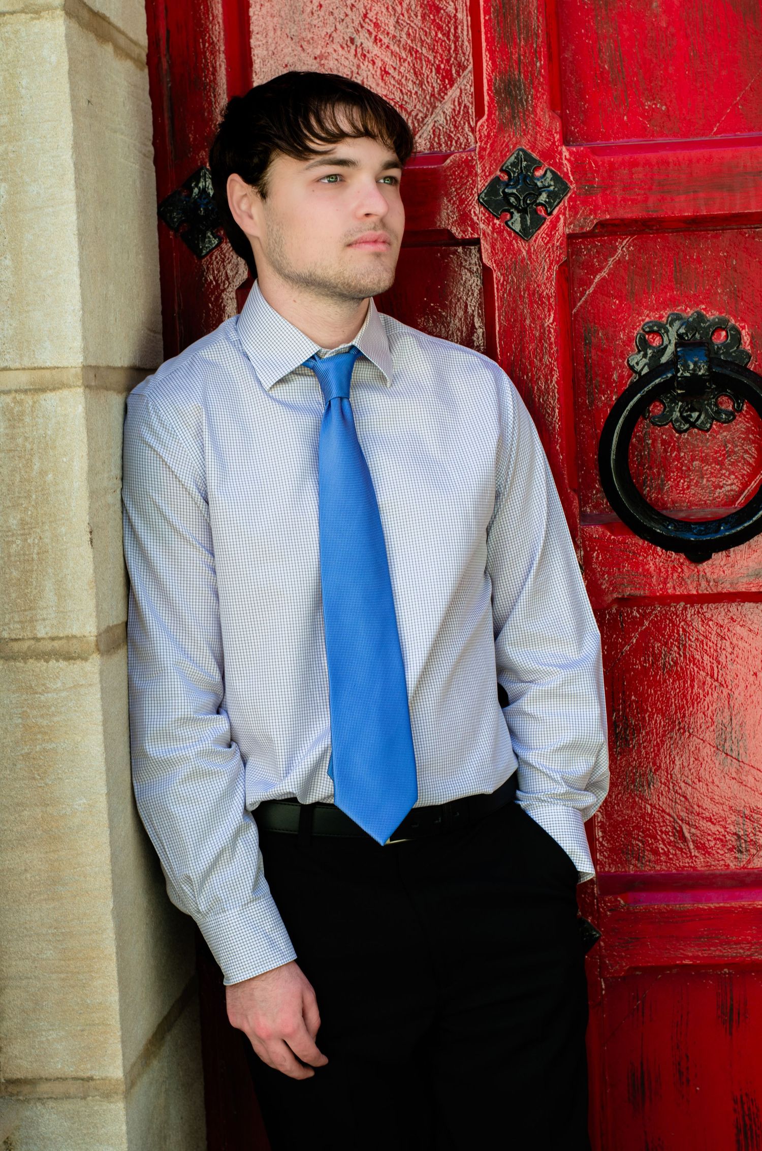 Young man in blue tie and gray dress shirt leaning against a rustic red door with ornate black knockers.