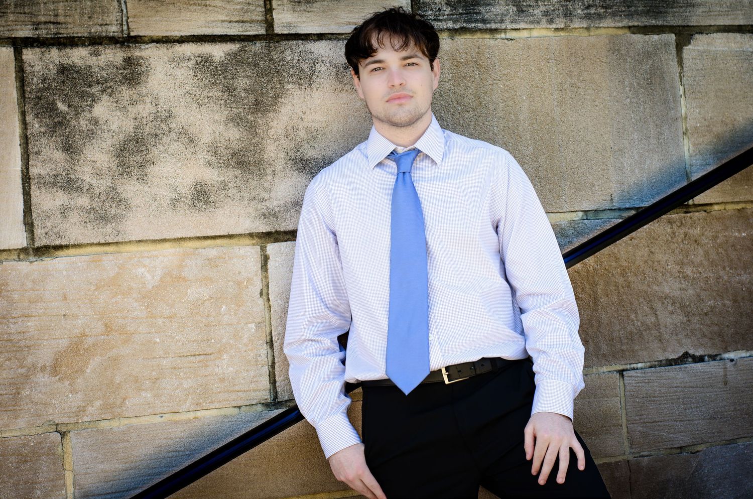 Young man in light blue dress shirt and tie posing against stone wall outdoors.