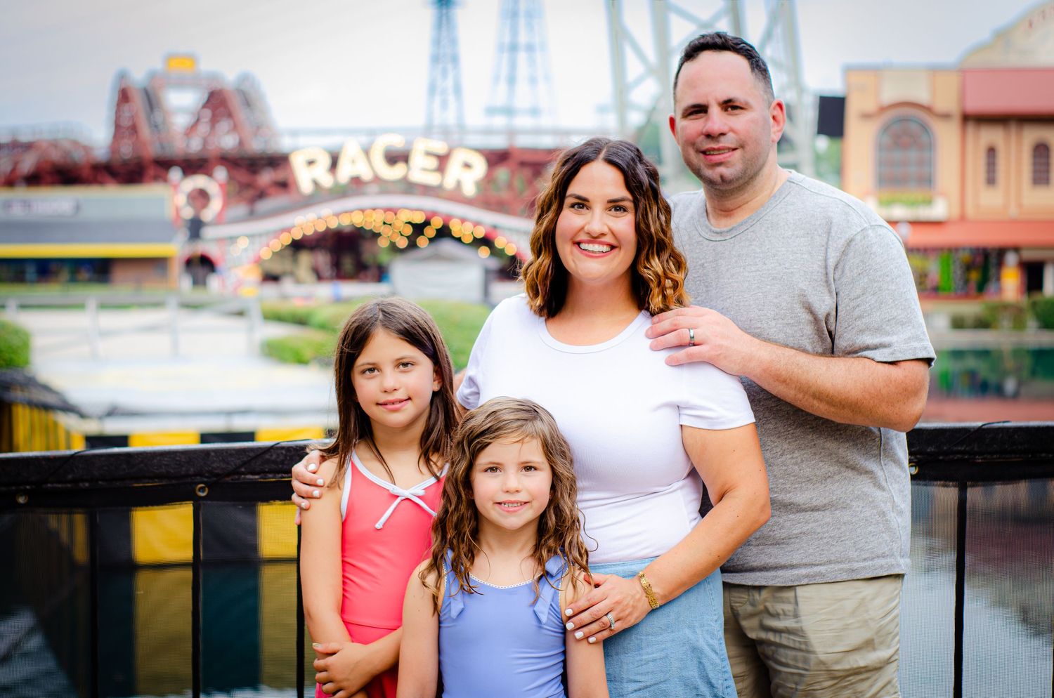 Family of four smiling at an amusement park with a Racer roller coaster sign in the background.