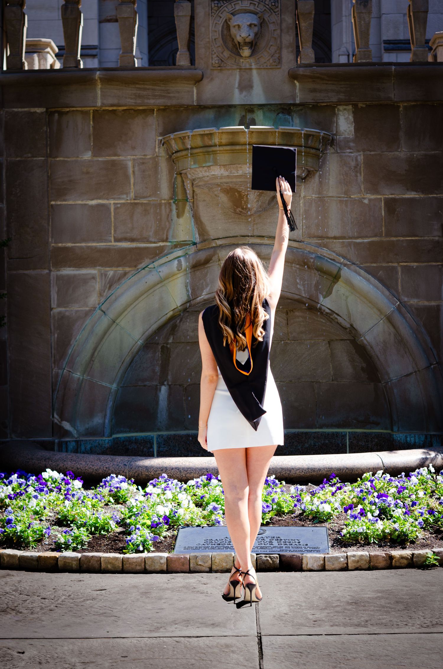 A female graduate raises her cap triumphantly in front of a stone fountain surrounded by purple flowers.