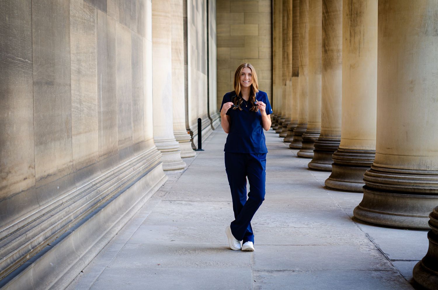 A woman in navy blue scrubs walks confidently through a classical stone colonnade.