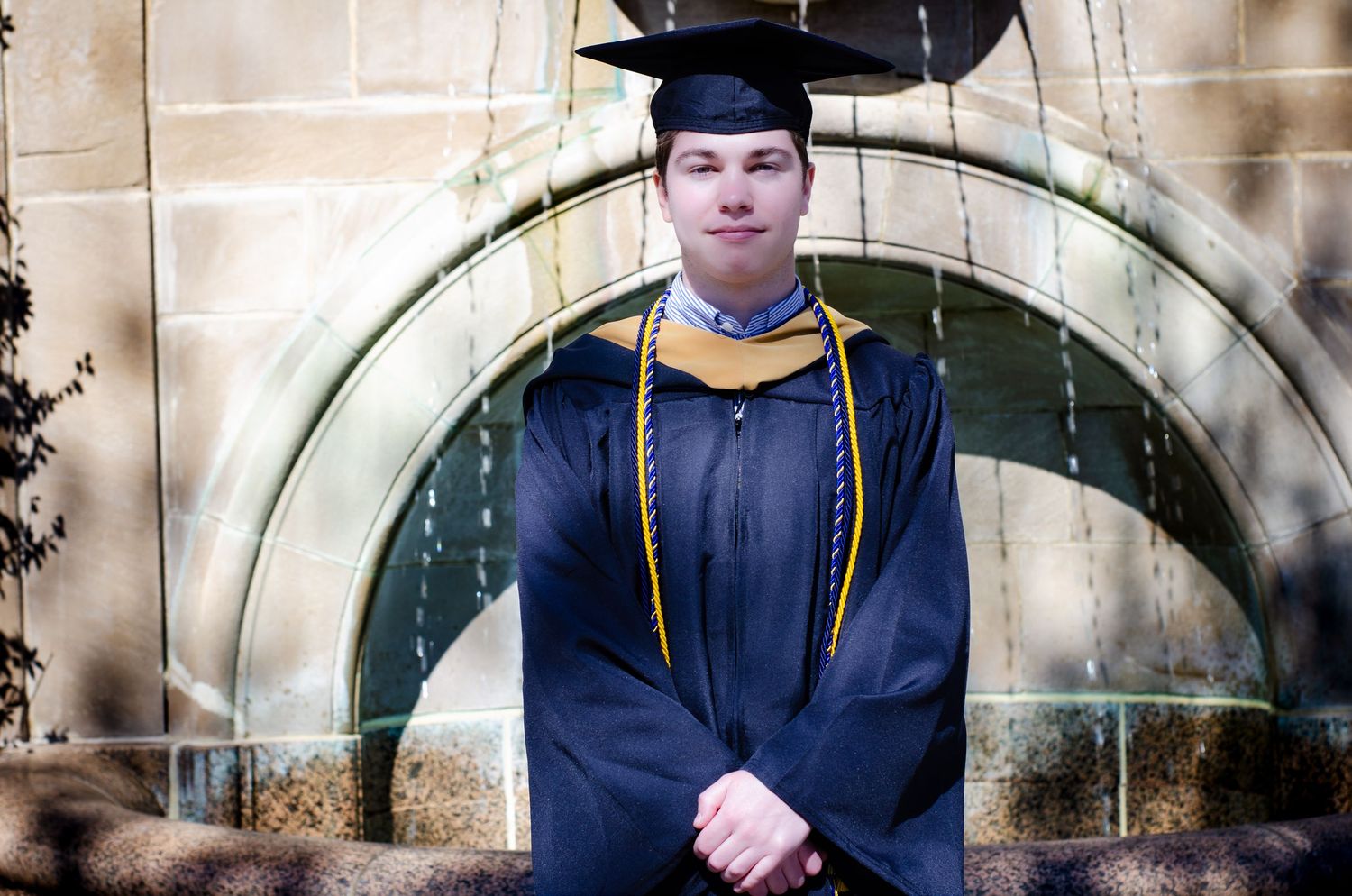 Graduate in black cap and gown with gold honor cords standing before stone arch fountain.