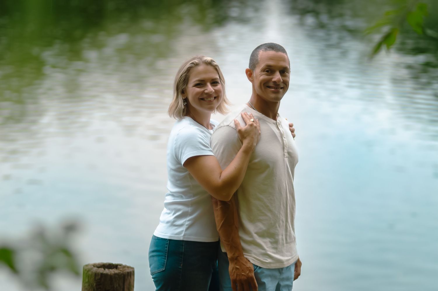 A couple stands together by a tranquil lake surrounded by lush greenery during a romantic photoshoot.