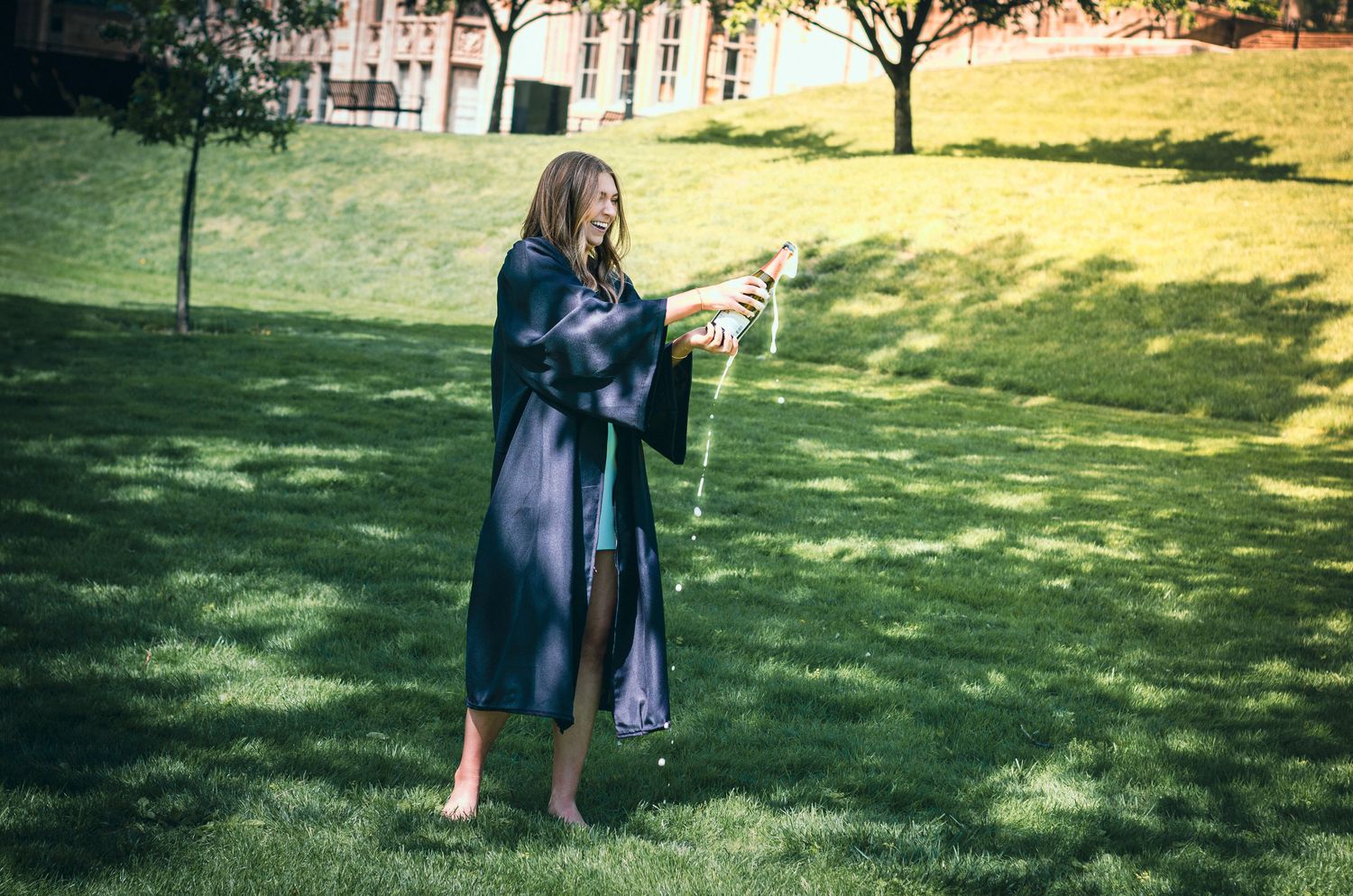 Graduate in black gown pops champagne bottle barefoot on green grass at outdoor college campus ceremony.