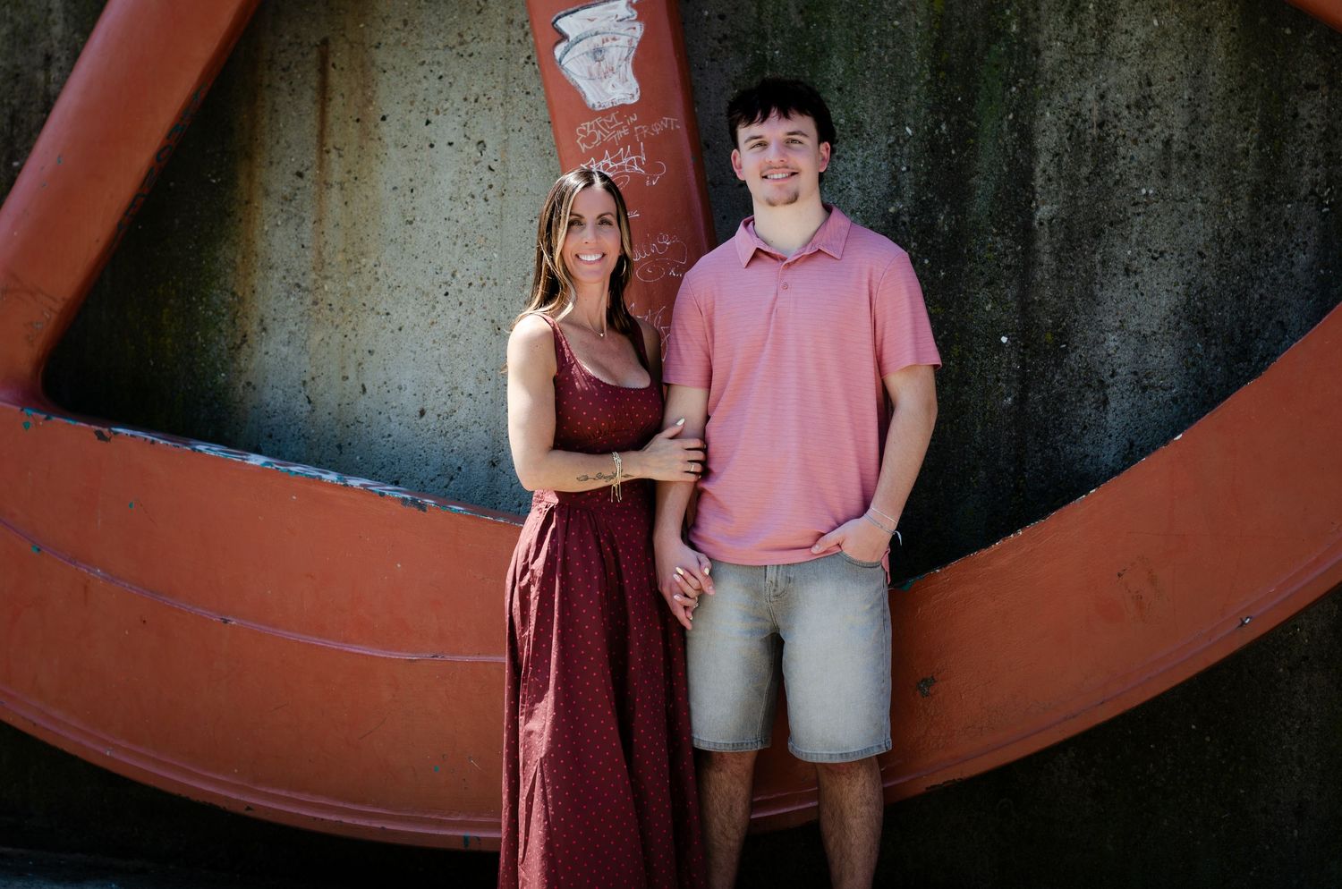 A smiling woman in a burgundy maxi dress poses with a tall young man in a pink polo shirt outdoors.