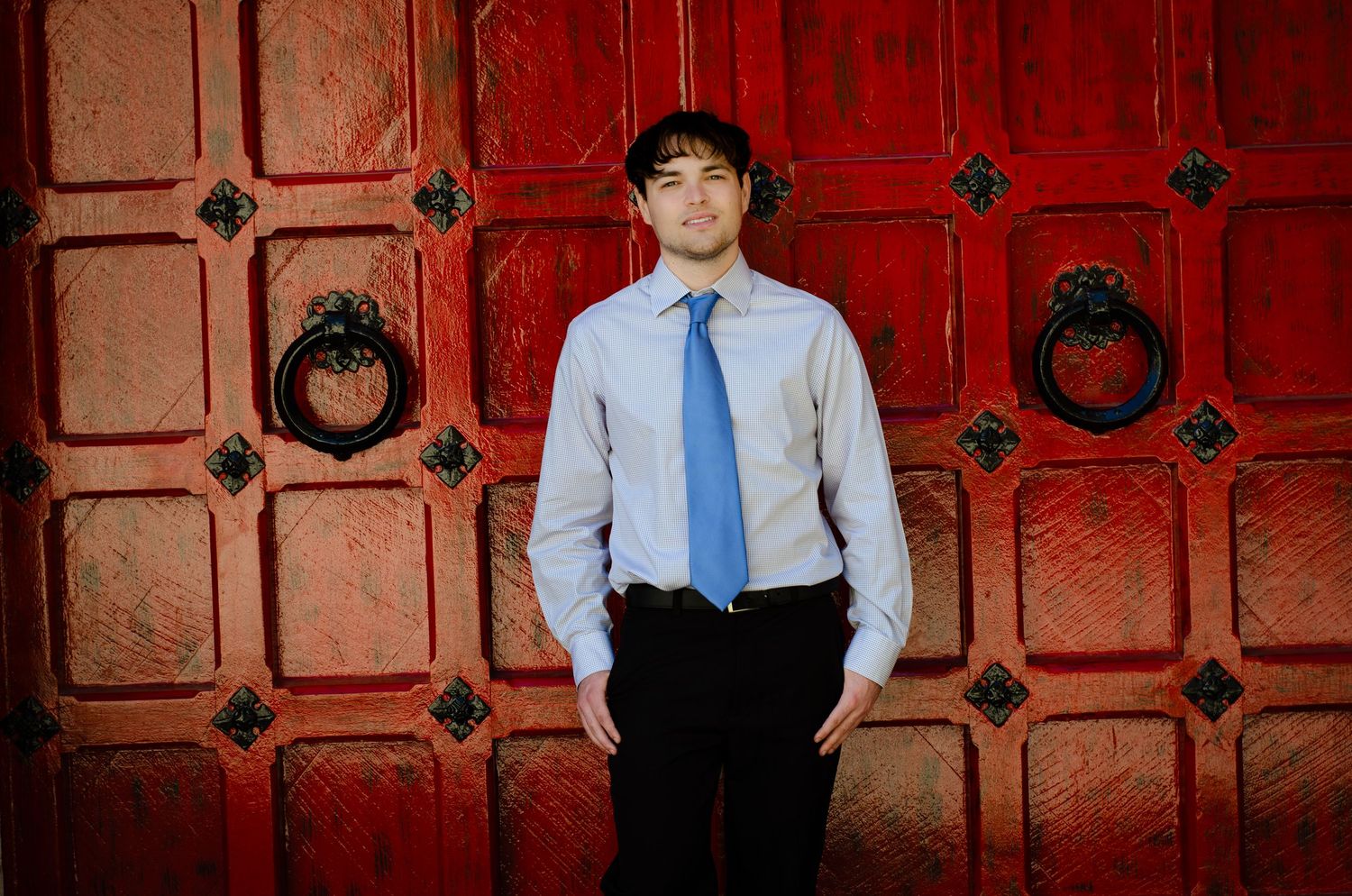 Young man in blue shirt and tie standing before ornate red doors with decorative iron knockers.