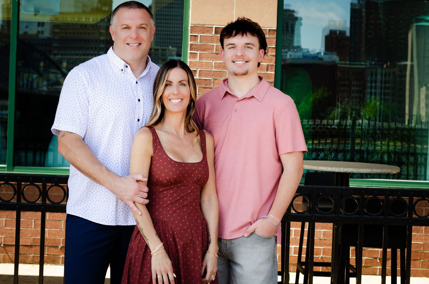 Three smiling people pose together outdoors near a brick building on a sunny day.