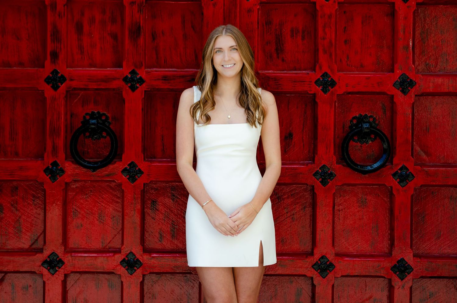 Young woman in white mini dress posing in front of ornate red doors with lion head knockers.