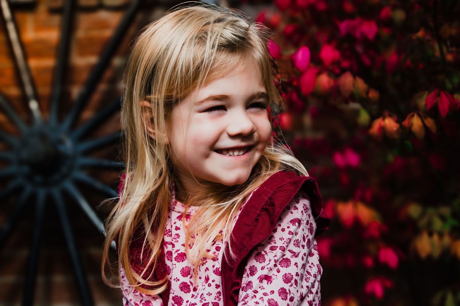 Young child wearing pink and burgundy outfit smiles joyfully against vibrant red autumn foliage background.