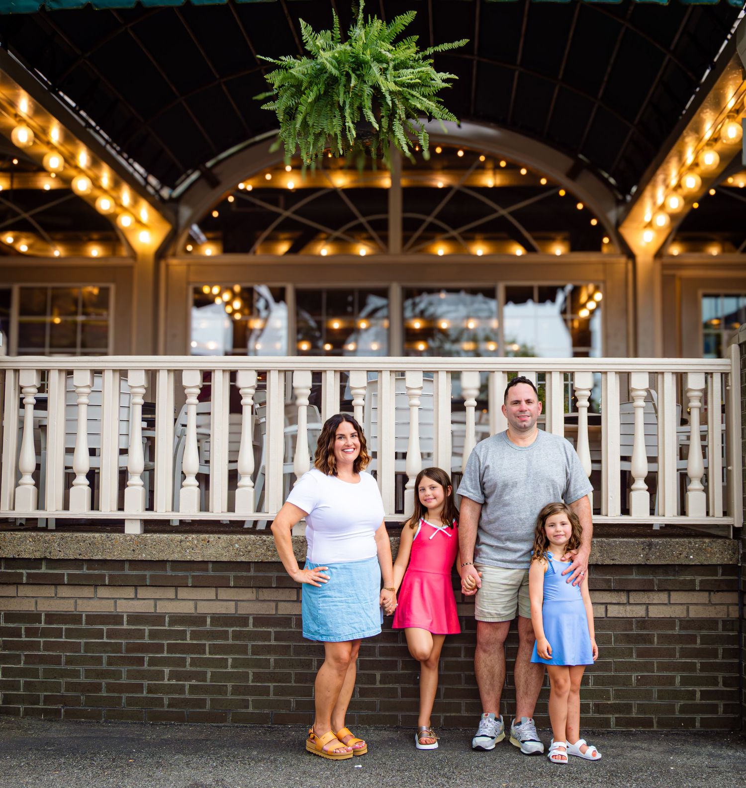 Family of four posing in front of a charming lit porch with string lights and a fern hanging basket.