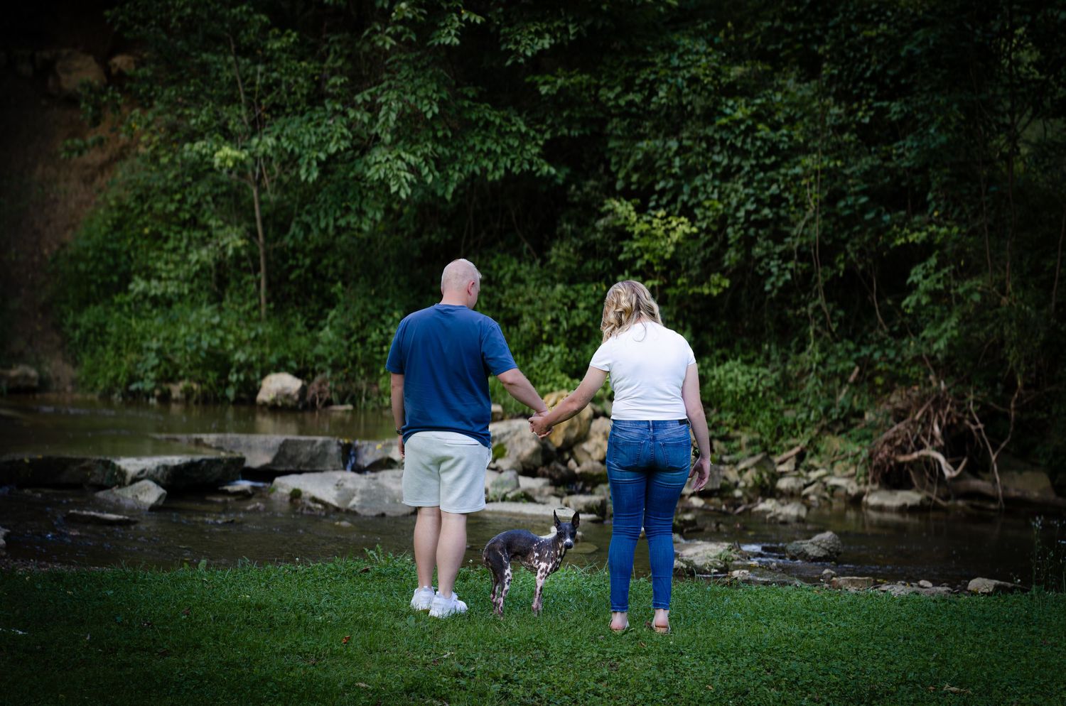 Couple with dog standing by a creek surrounded by lush green trees in a park setting.