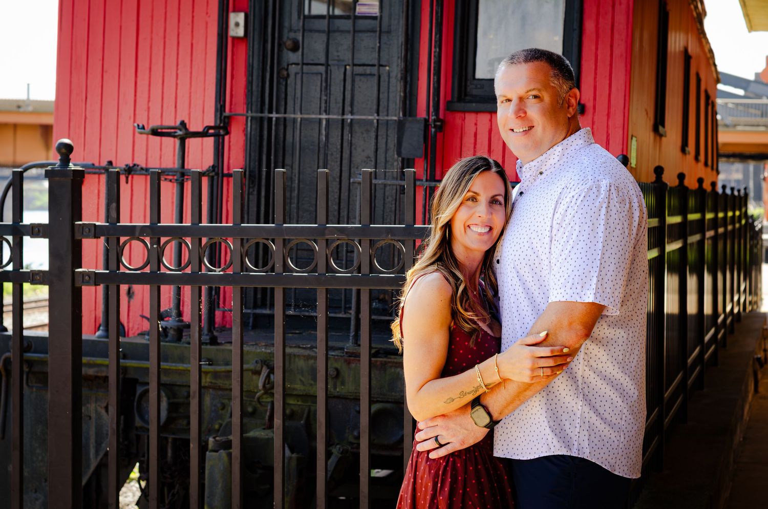 A happy couple embraces in front of a red building with black iron railings outdoors.