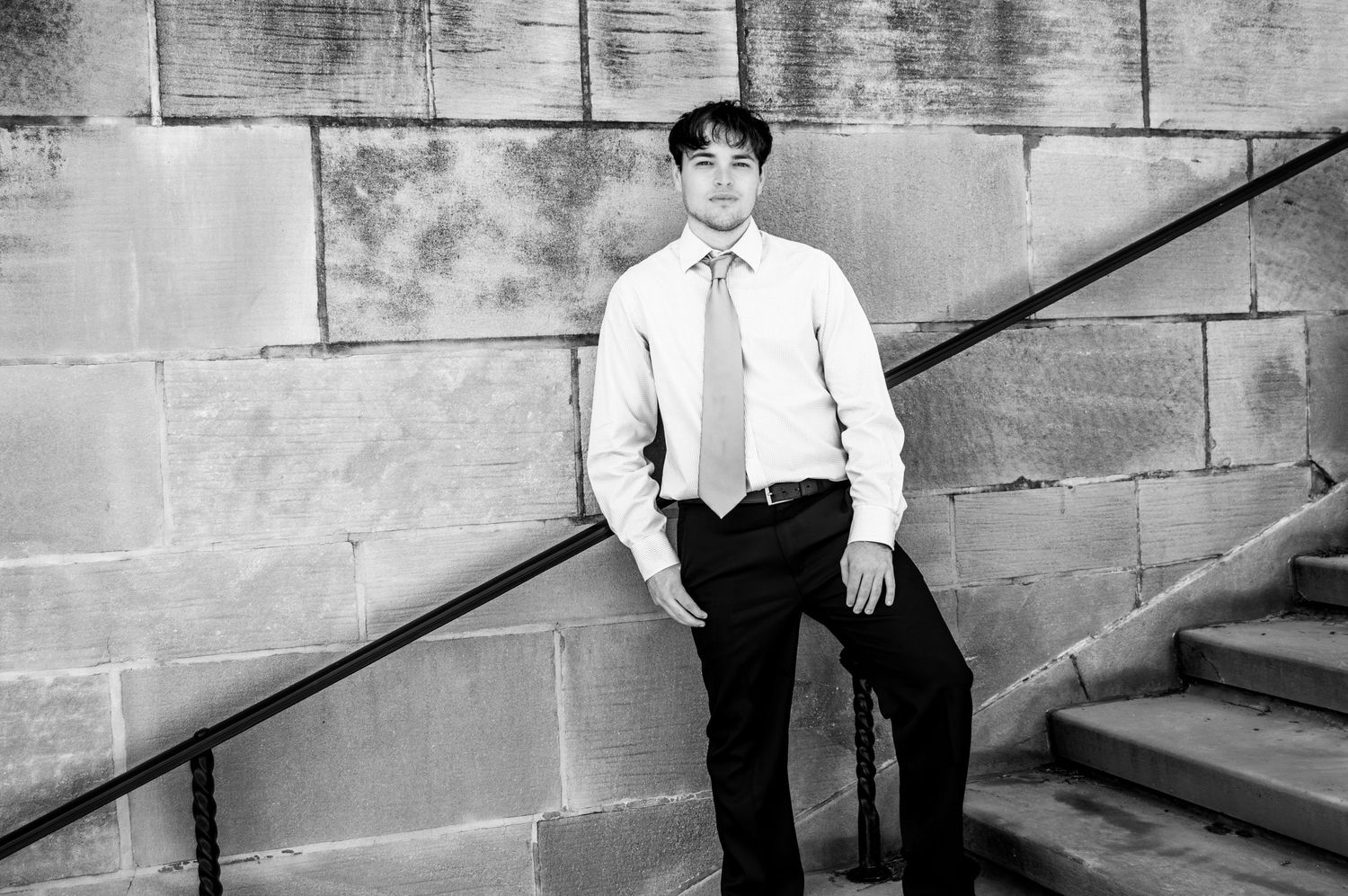 Black and white portrait of young man in dress shirt and tie leaning against stone wall by stairs.
