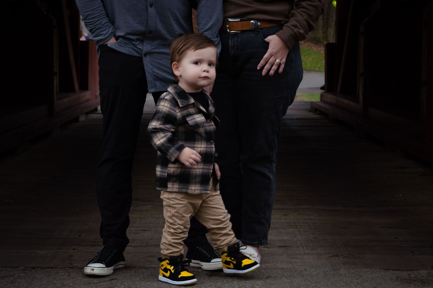 Small child stands in a plaid jacket and khaki pants wearing colorful sneakers in a dark covered walkway.