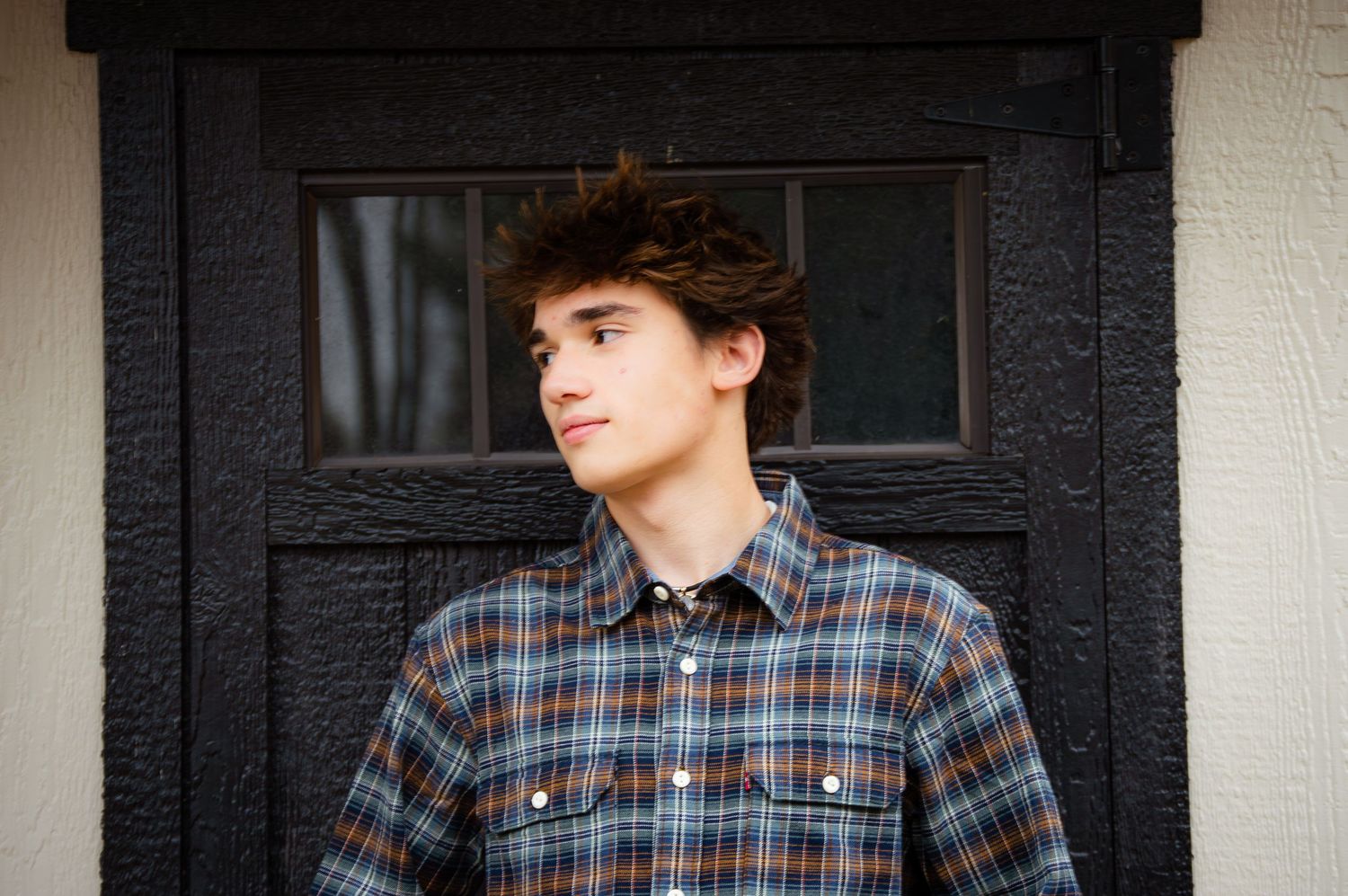 Person in plaid shirt and dark pants stands against white columns and black shutters.