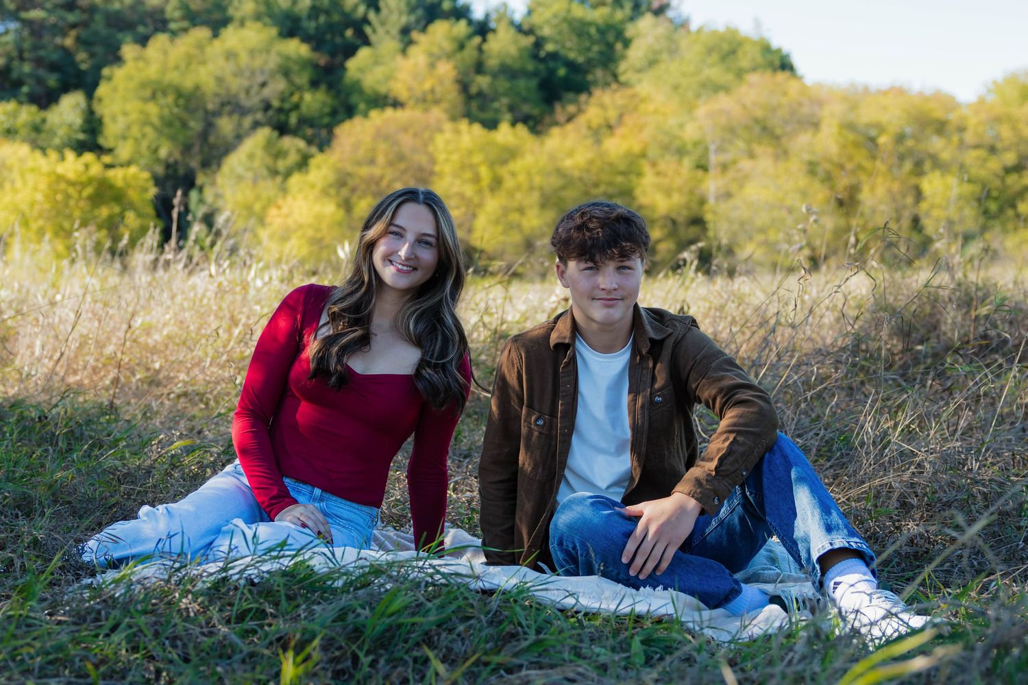 Two teenagers sitting on a blanket in a golden autumn field, smiling at the camera.