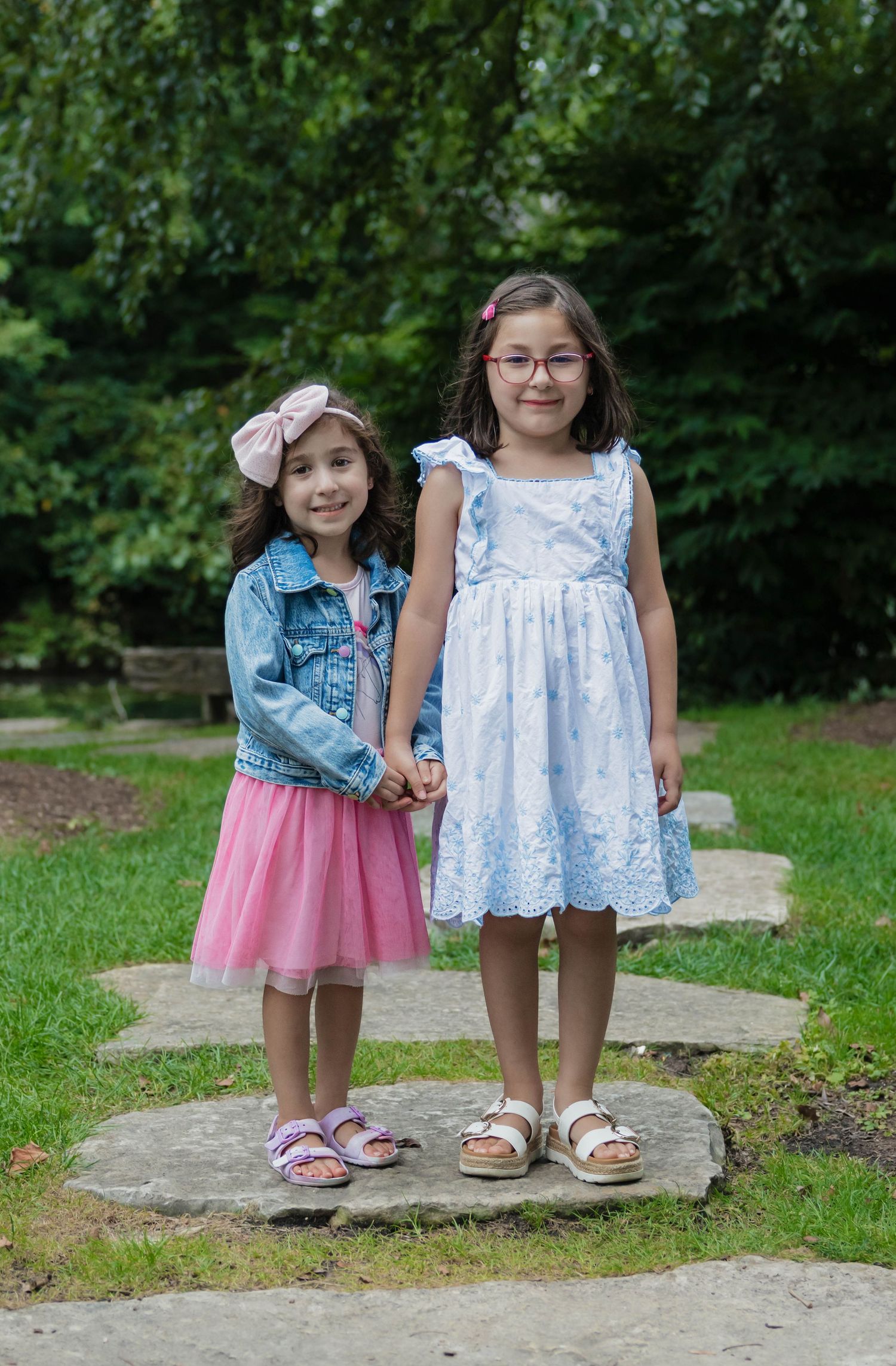 Two young girls holding hands on stepping stones in a green garden, one in pink tutu, one in blue dress.