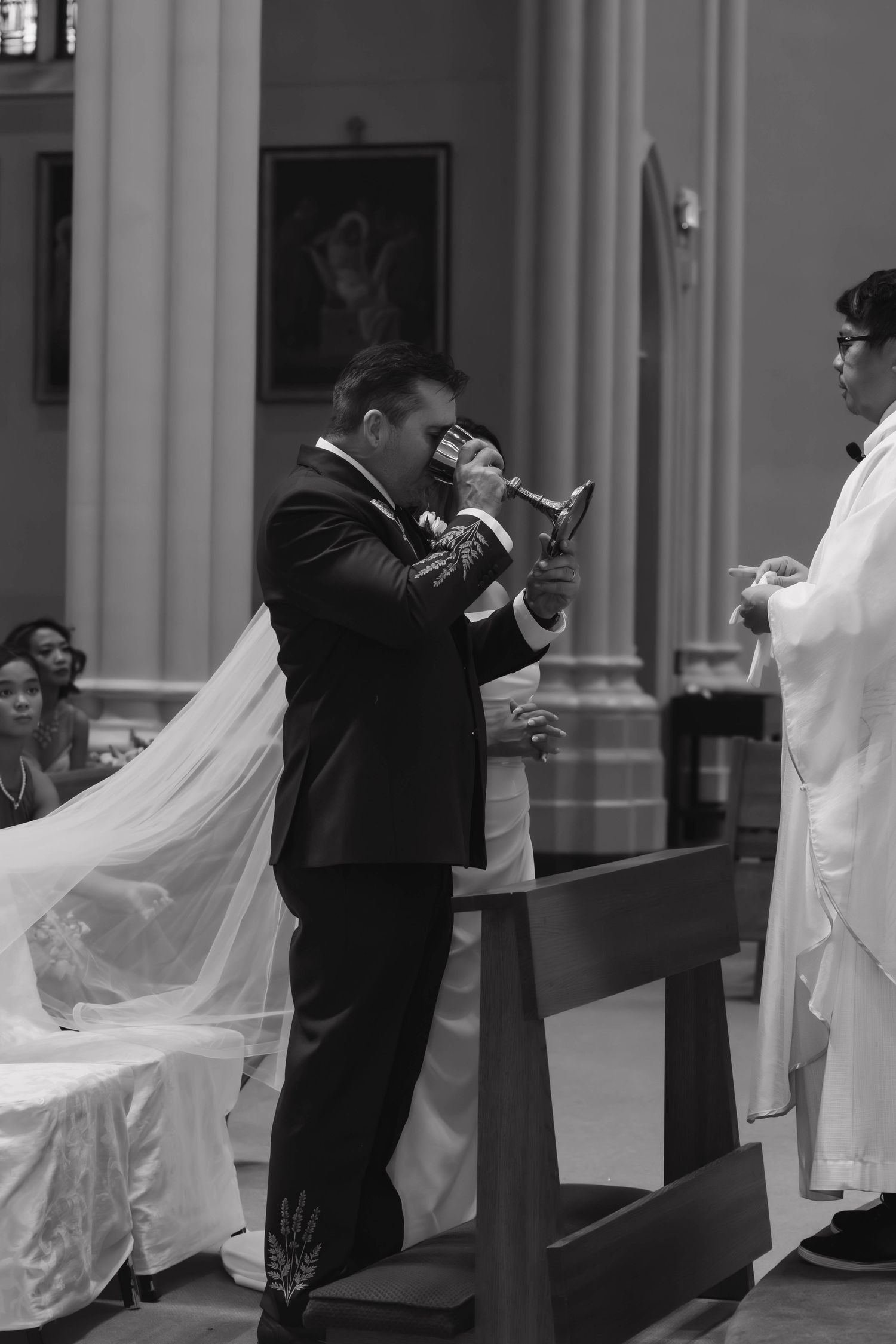 Groom in black suit receives communion from priest during black and white wedding ceremony photo.