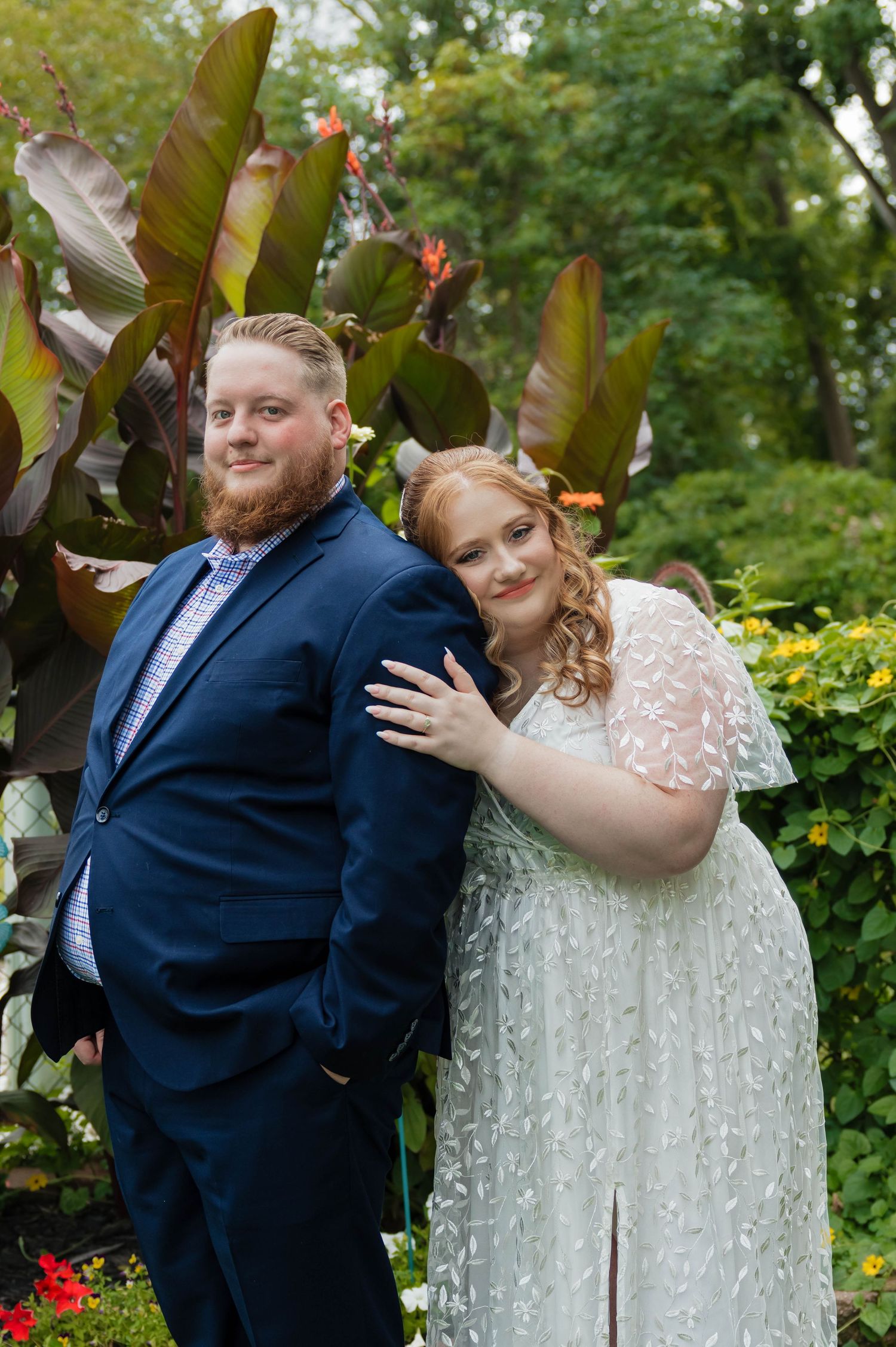 Wedding couple posing in garden, bride in white lace gown leaning on groom in navy suit.