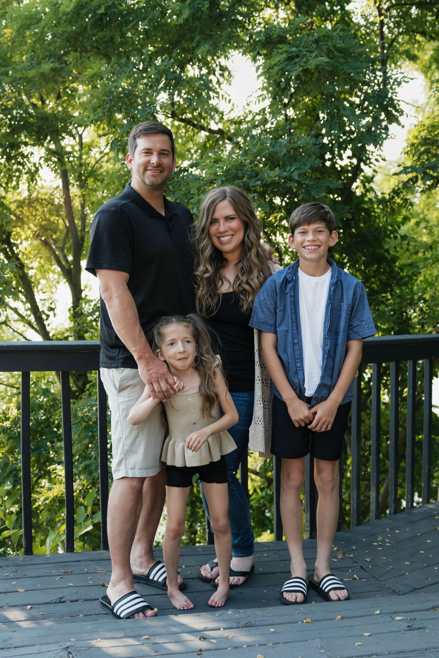 Happy family of four posing on a wooden deck with green trees in the background.