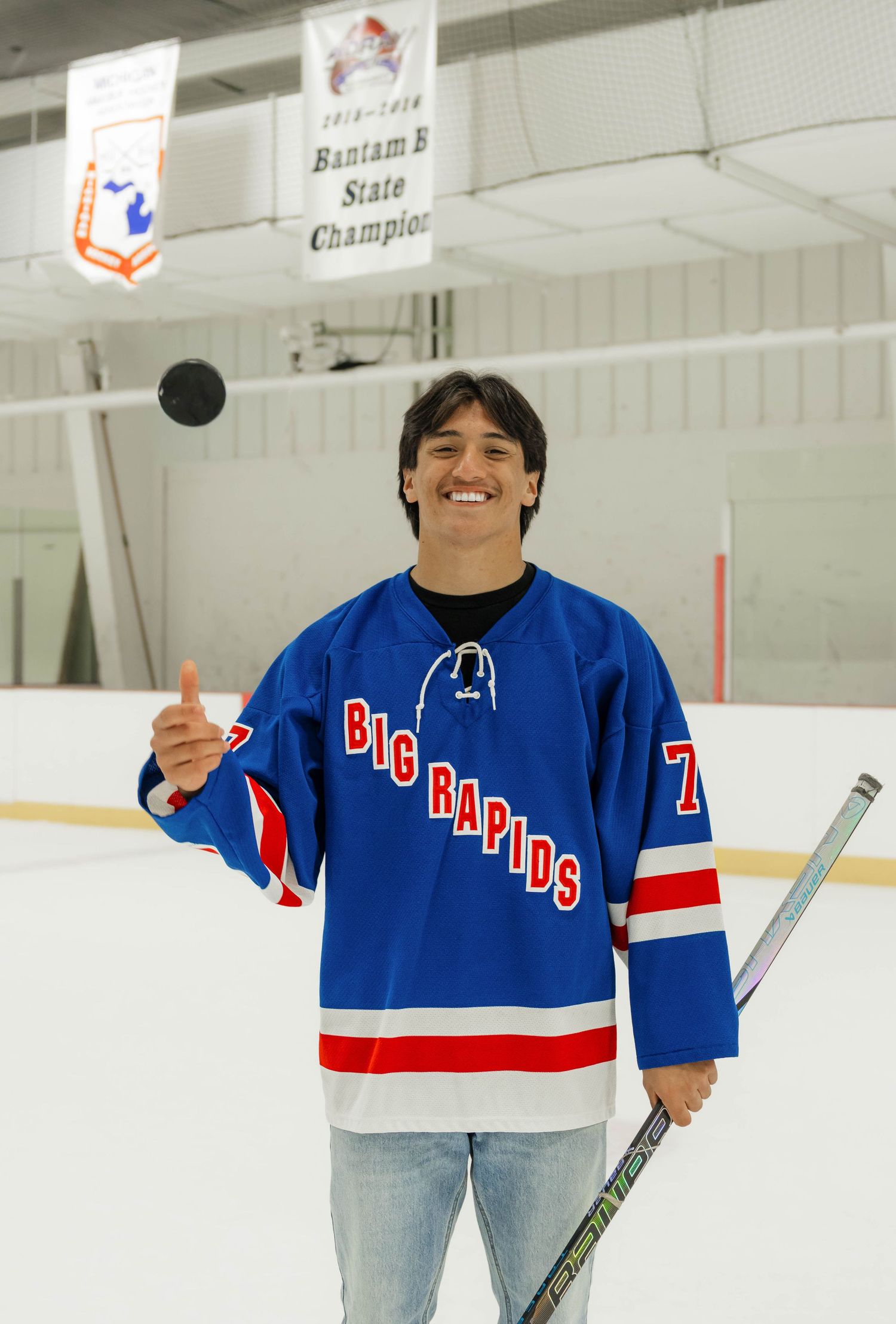 Smiling teen in Big Rapids blue hockey jersey tosses puck on ice rink.