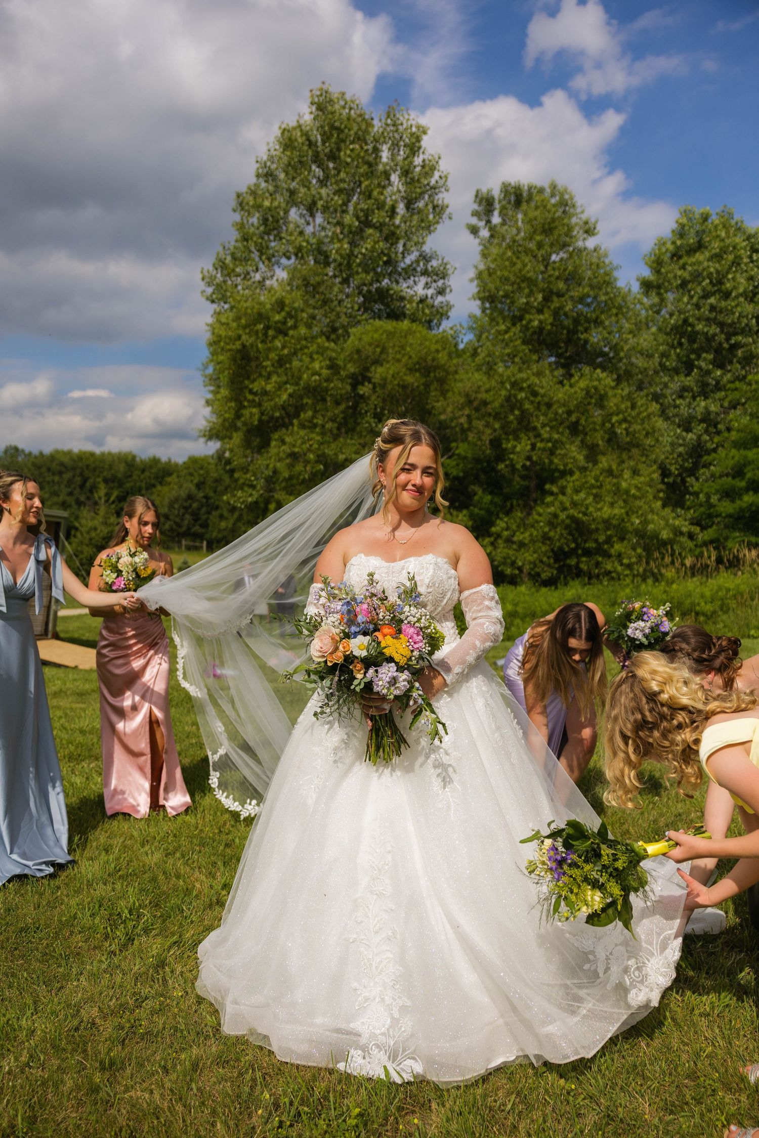 Bride in white ballgown holds colorful bouquet outdoors as bridesmaids in pink and blue adjust her veil and dress.