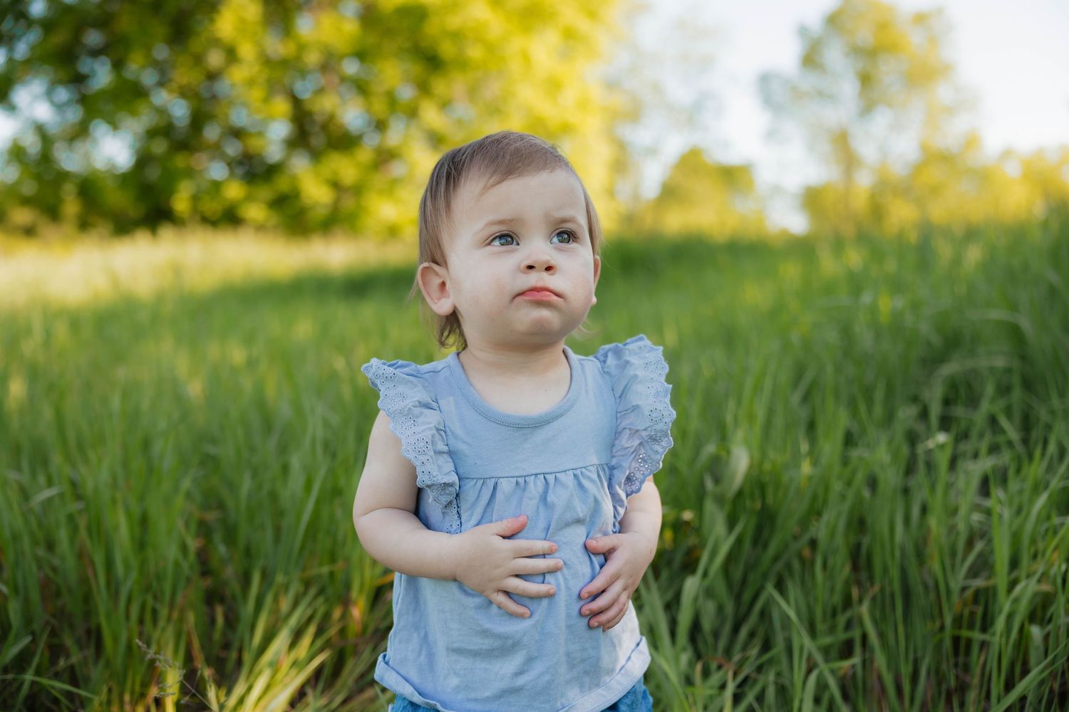 Cute toddler girl in a blue ruffle top standing in a vibrant green field on a sunny day.