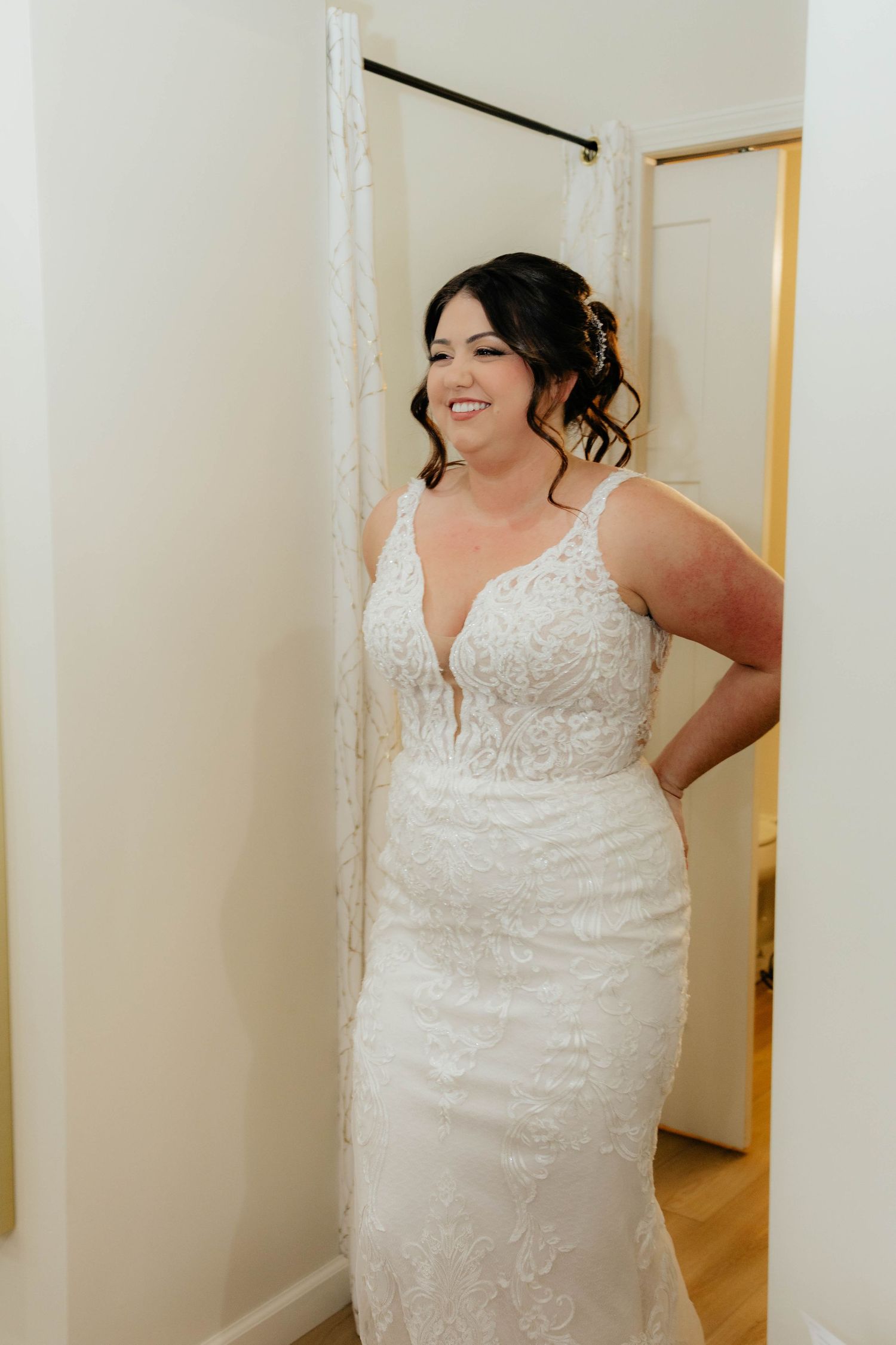 Smiling bride in fitted lace V-neck wedding gown standing in a dressing room doorway.