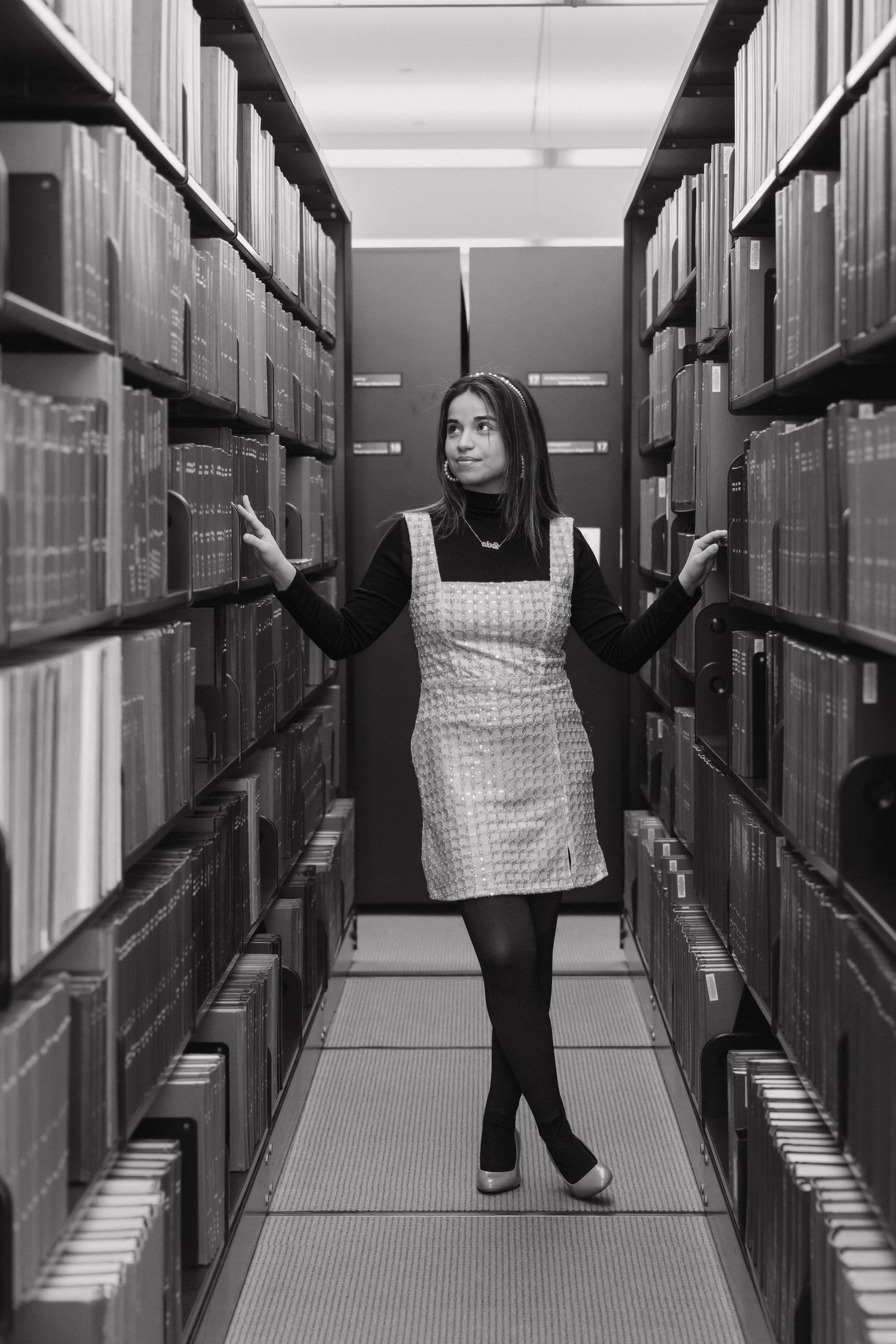 Black and white photo of a woman browsing between tall library bookshelves in a white dress.