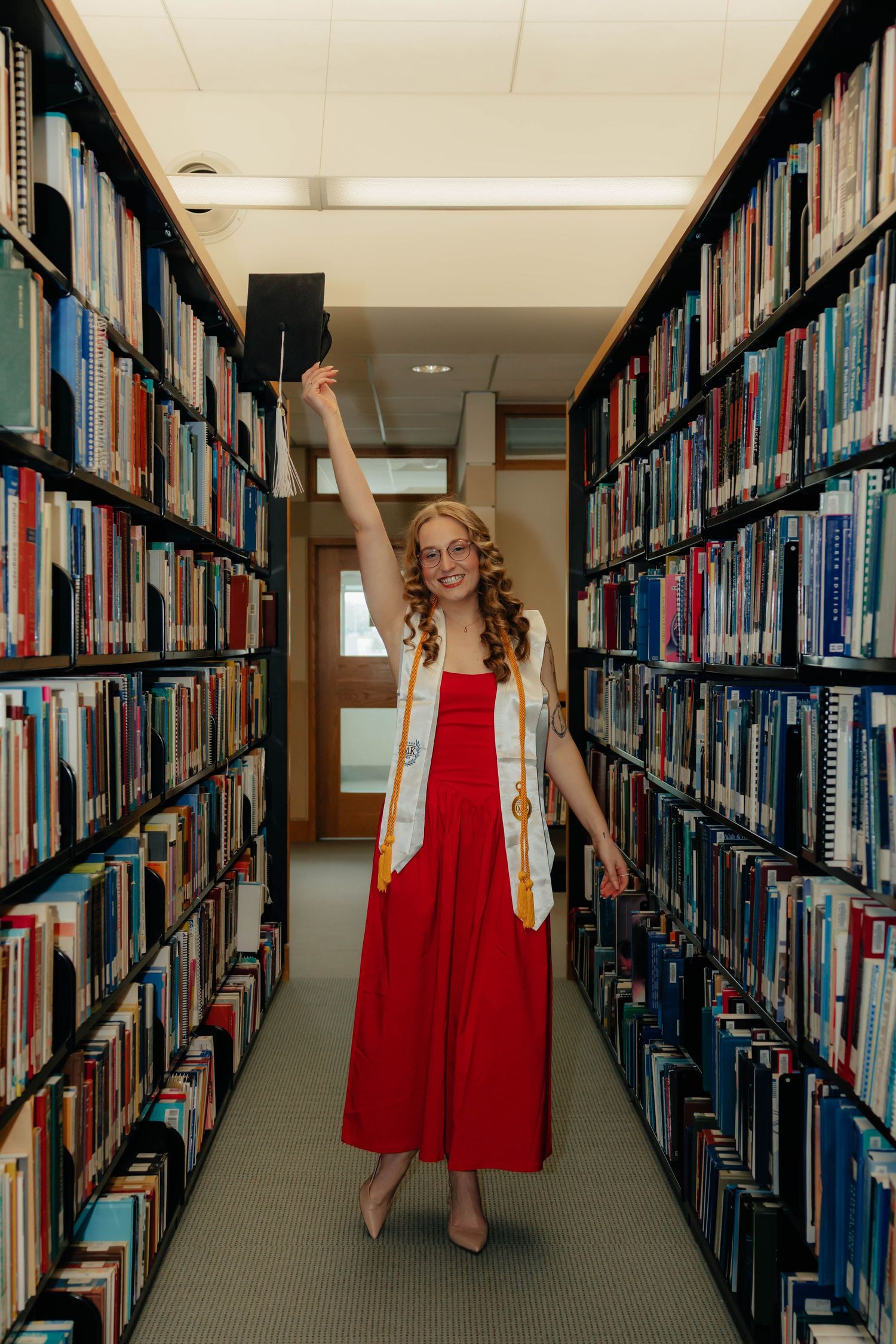 A smiling graduate in a red dress holds her cap up in a library aisle lined with colorful books.