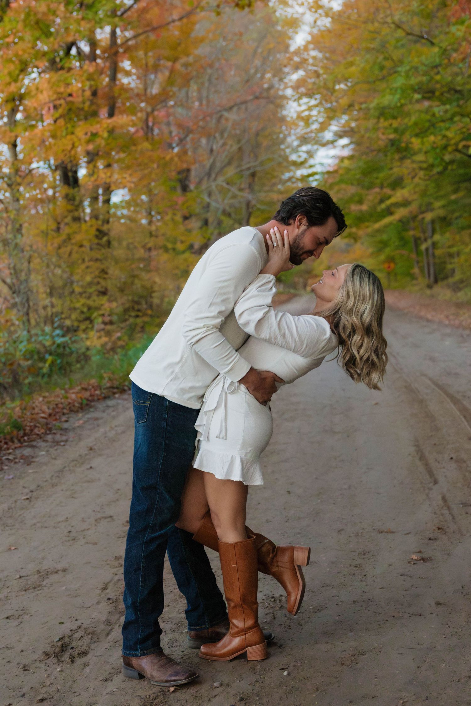 Couple sharing a passionate dip kiss on a scenic fall dirt road lined with vibrant autumn trees.