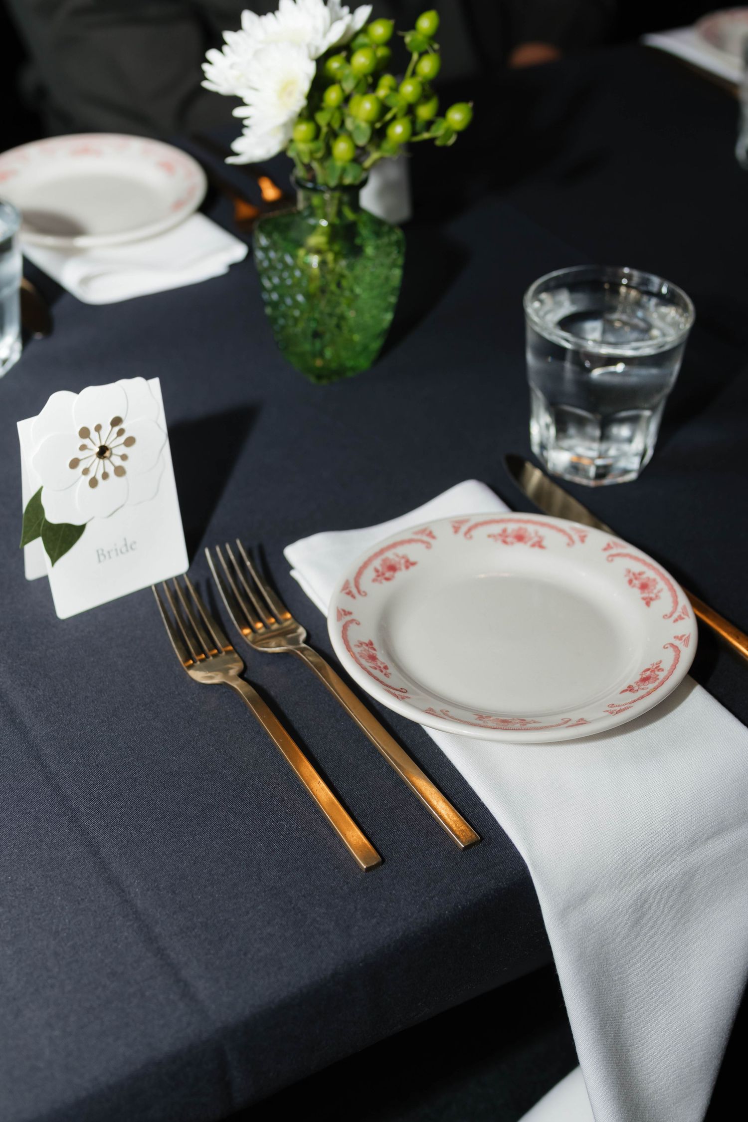 Formal wedding reception place setting with gold cutlery, floral plate, white napkin, and flower centerpiece.