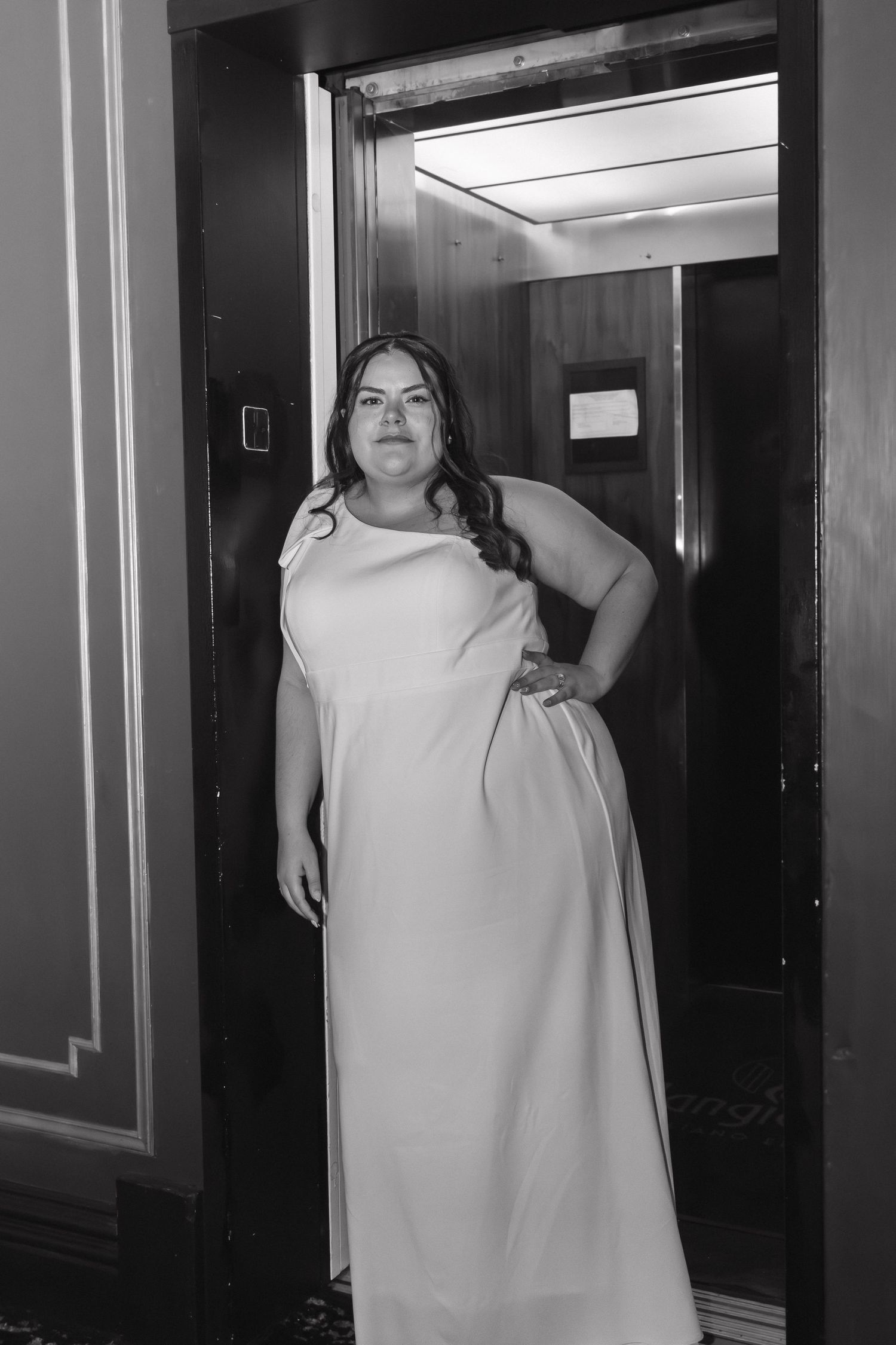 Bride in elegant white gown posing in elevator doorway, black and white wedding photo.