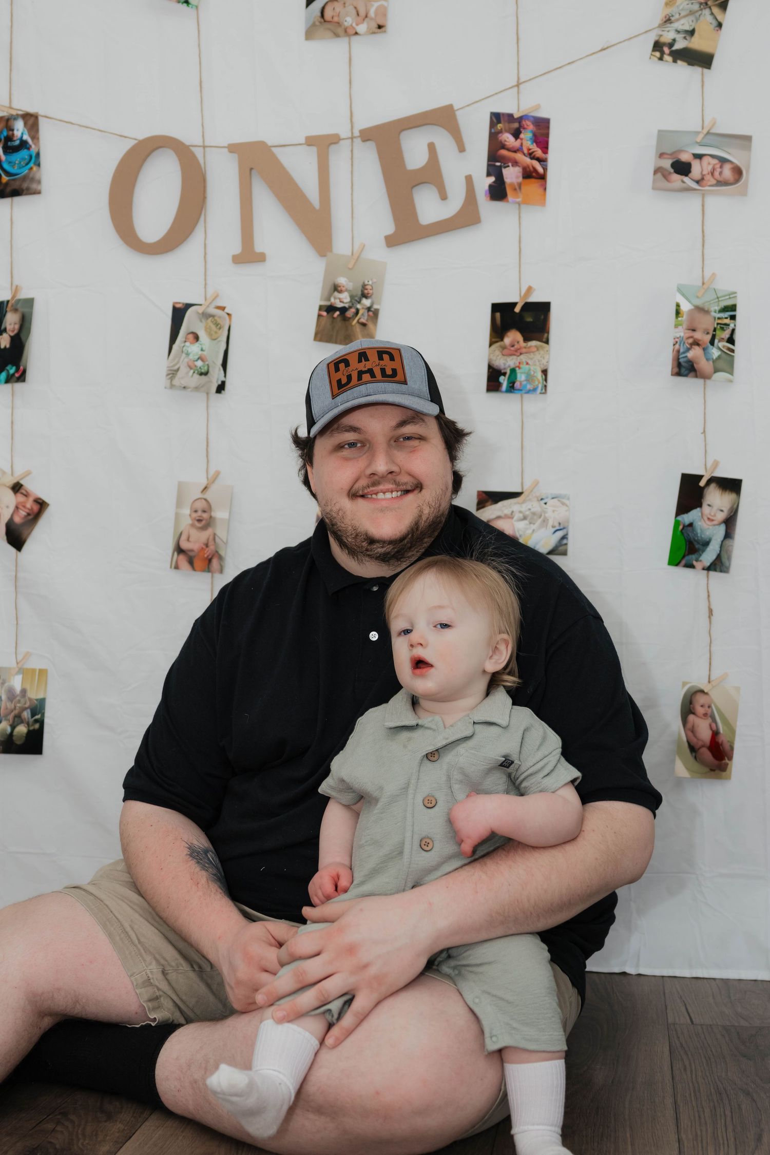 Father smiles holding one-year-old son in sage romper before ONE banner photo display wall.