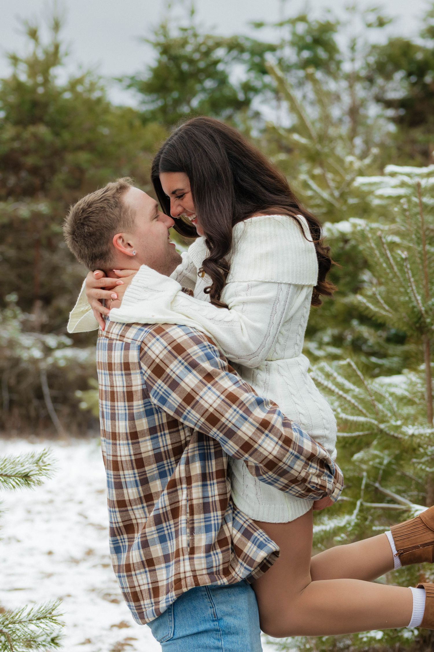 Couple embracing in snowy pine forest, man lifting woman in white sweater during winter engagement photo session.