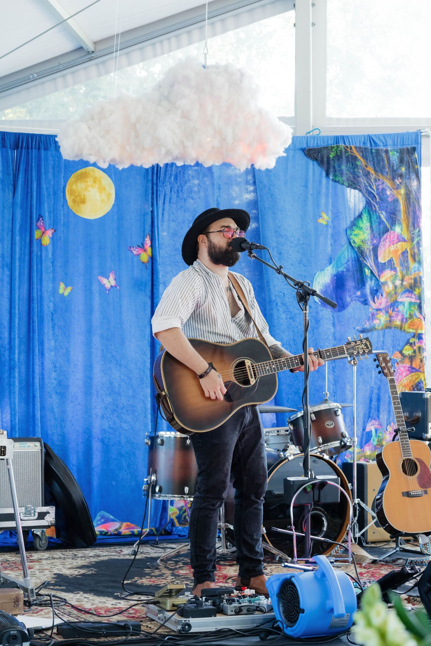 Bearded guitarist in black hat performs acoustic set on decorated stage at outdoor wedding reception.