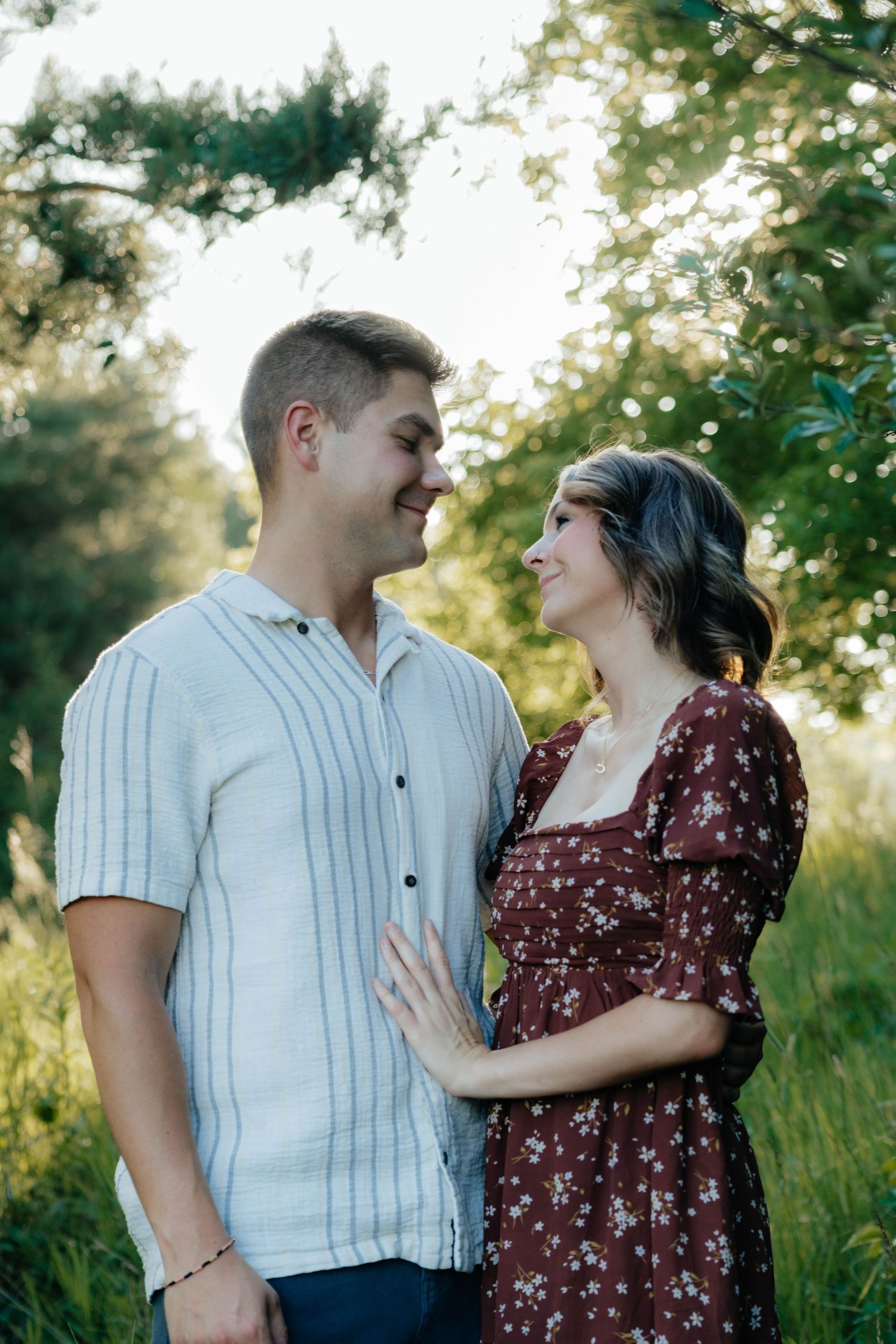 Smiling couple gazing at each other in a lush green outdoor setting, woman in floral dress, man in striped shirt.